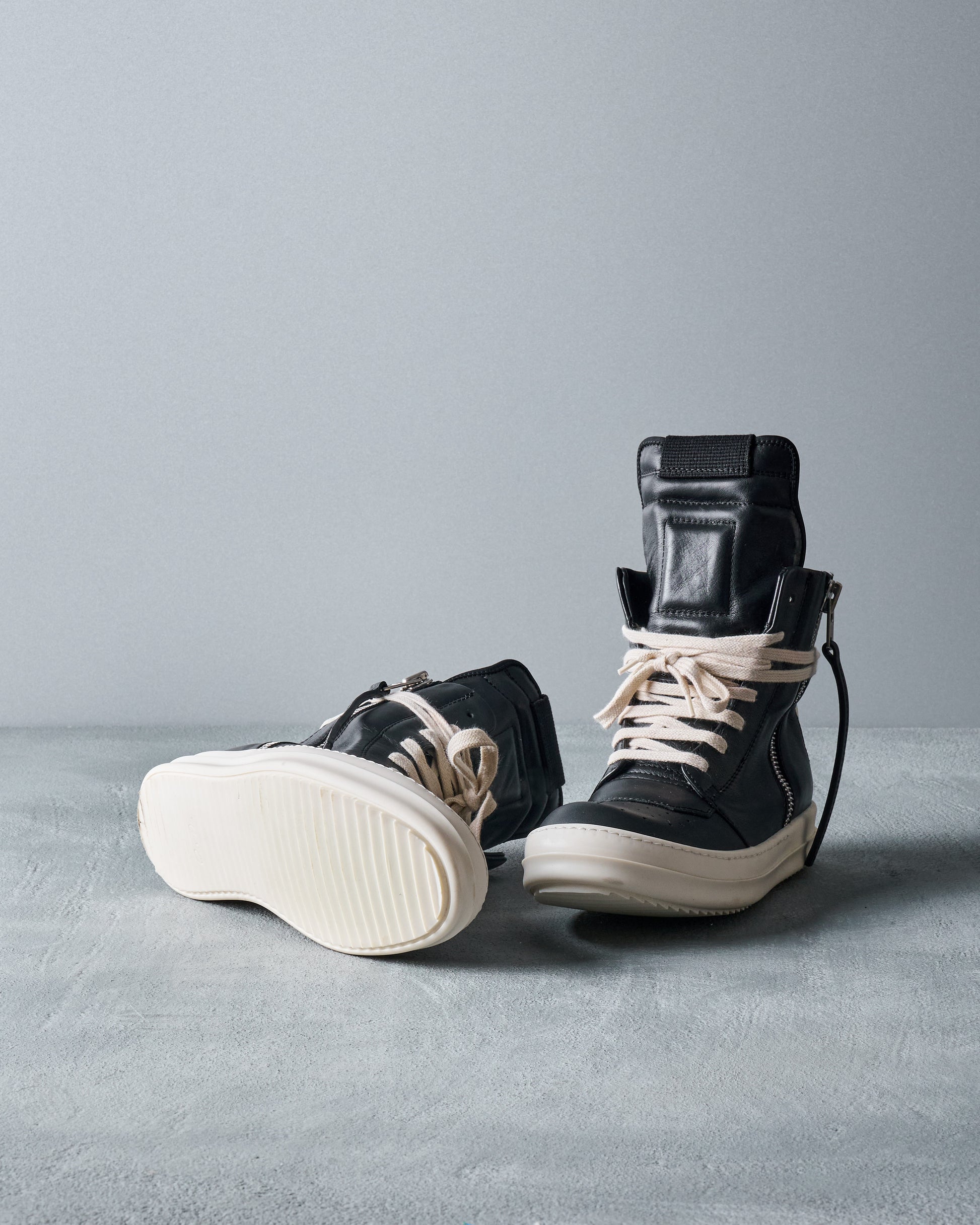 Pair of black high-top Rick Owens sneakers with white soles on a gray surface and background.