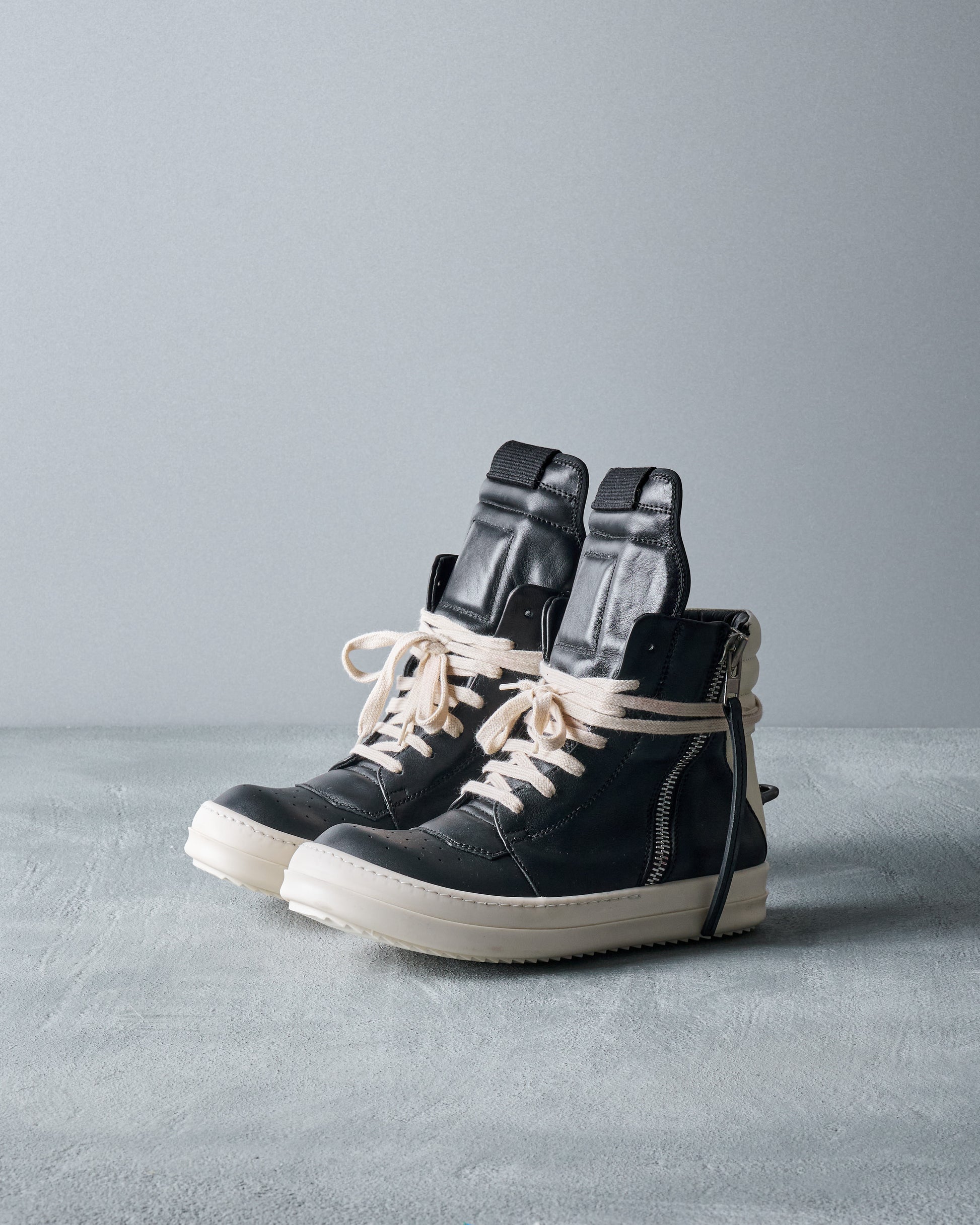 Black high-top Rick Owens sneakers with white laces on a gray surface and background