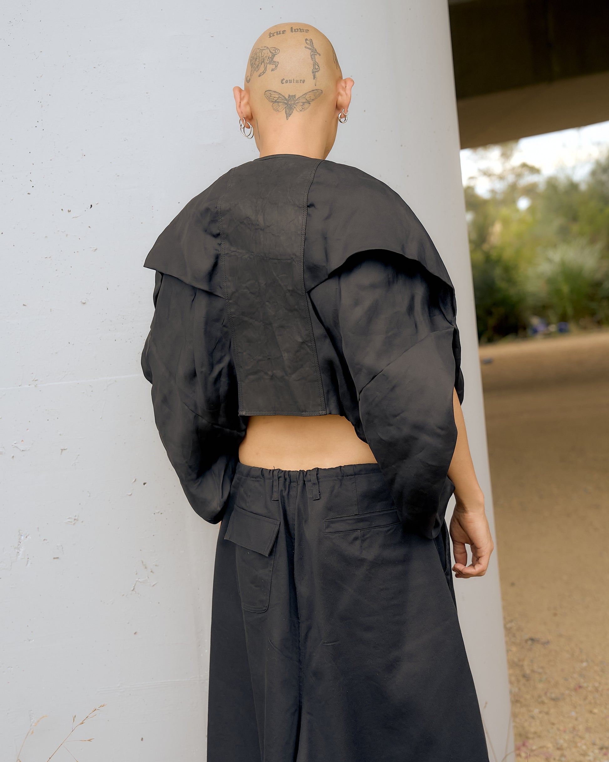 A person stands with their back to the camera, wearing an oversized Rick Owens black moto outfit. Tattoos are visible on their shaved head, set against an outdoor backdrop.