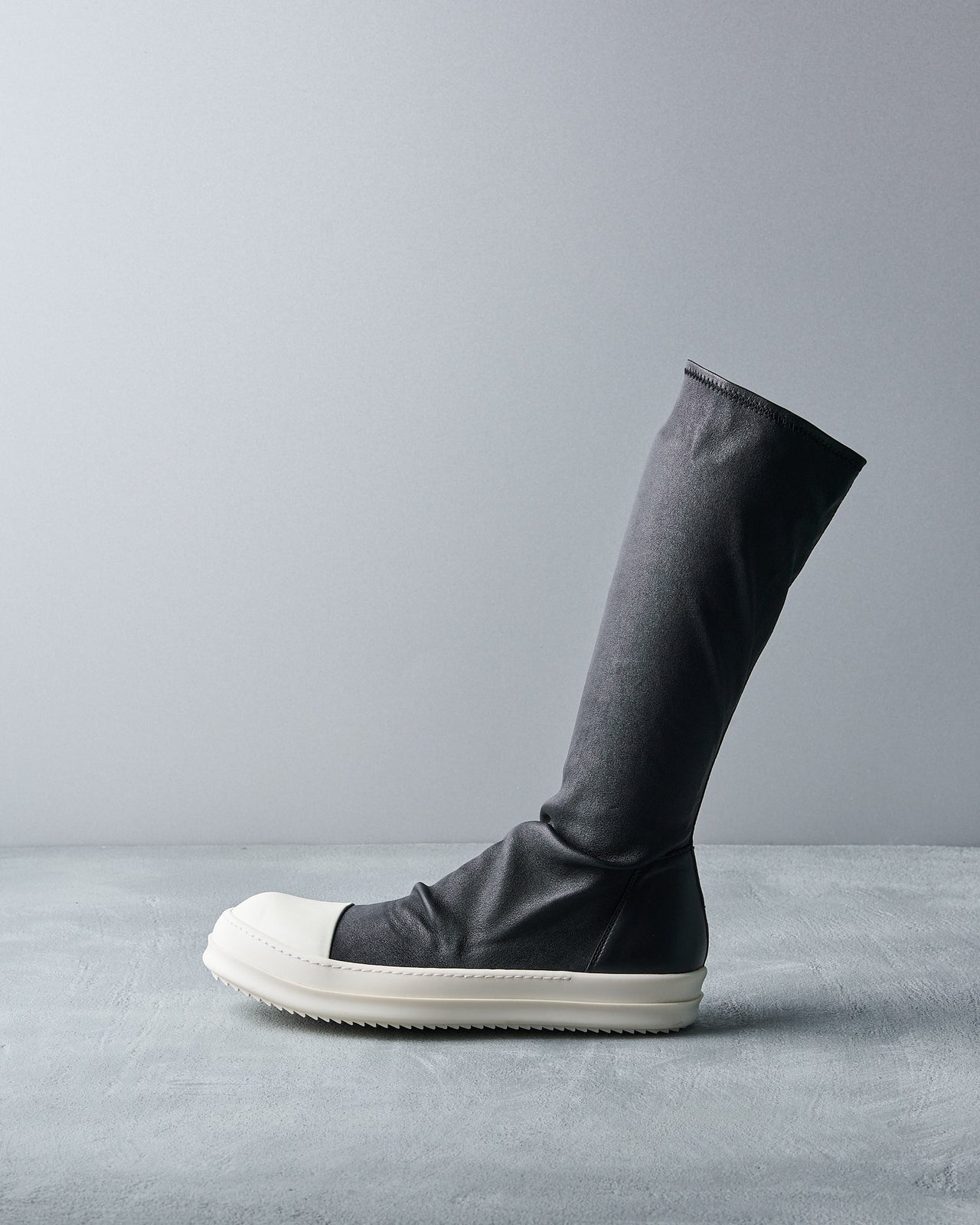 A sleek, high-top black Rick Owens boot with a white rubber-like sole and toe cap stands upright on a light gray surface, set against a matching gray backdrop.