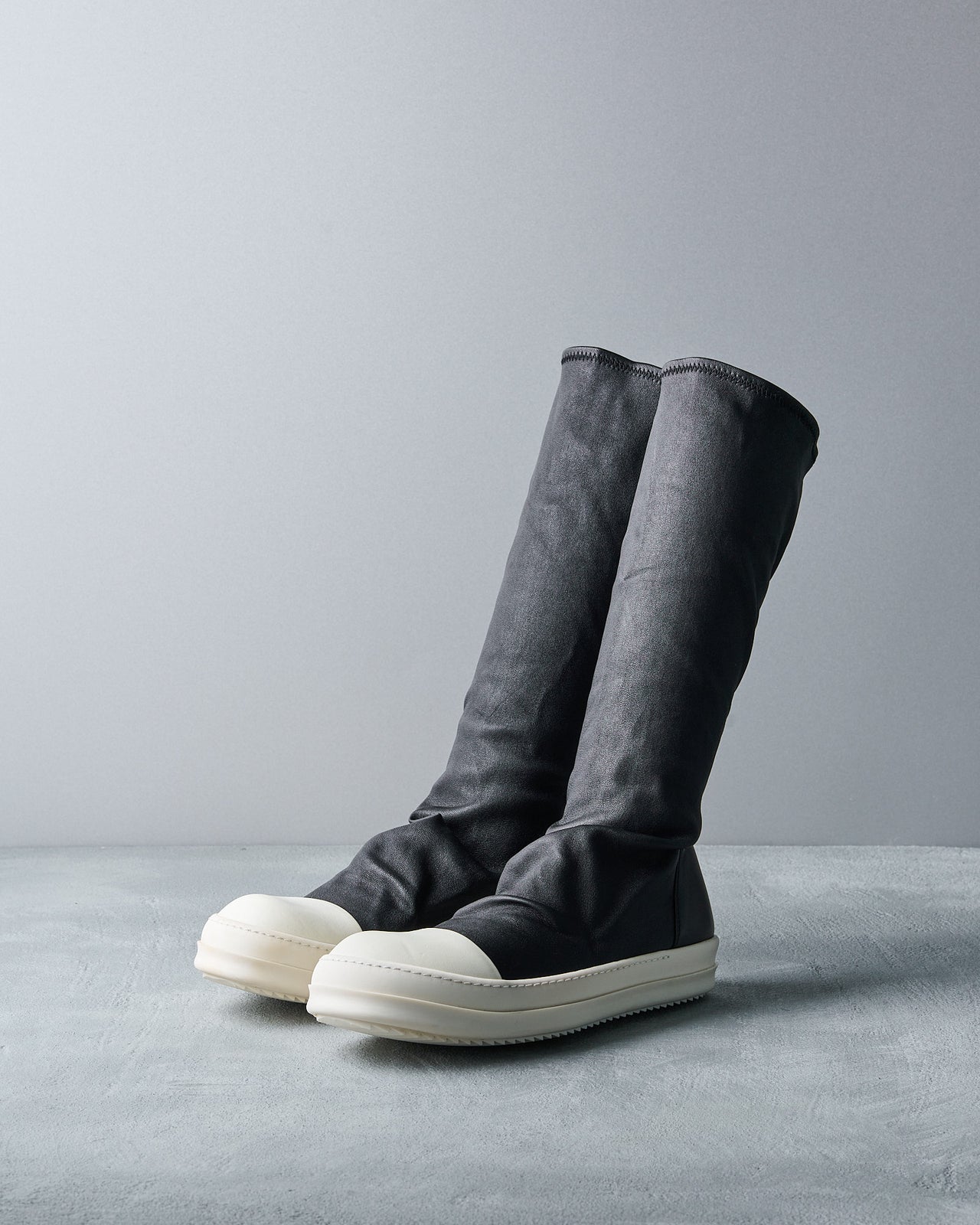 A sleek, high-top black Rick Owens boot with a white rubber-like sole and toe cap stands upright on a light gray surface, set against a matching gray backdrop.