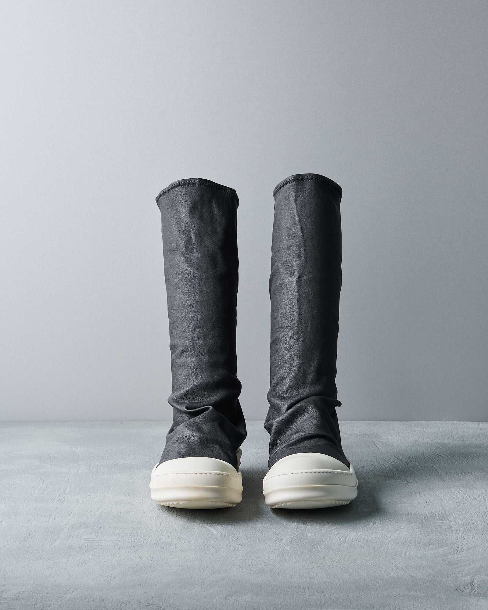 Tall, dark grey fabric Rick Owens boots with white rubber soles stand side by side on a light grey surface against a matching backdrop, conveying a minimalist style.