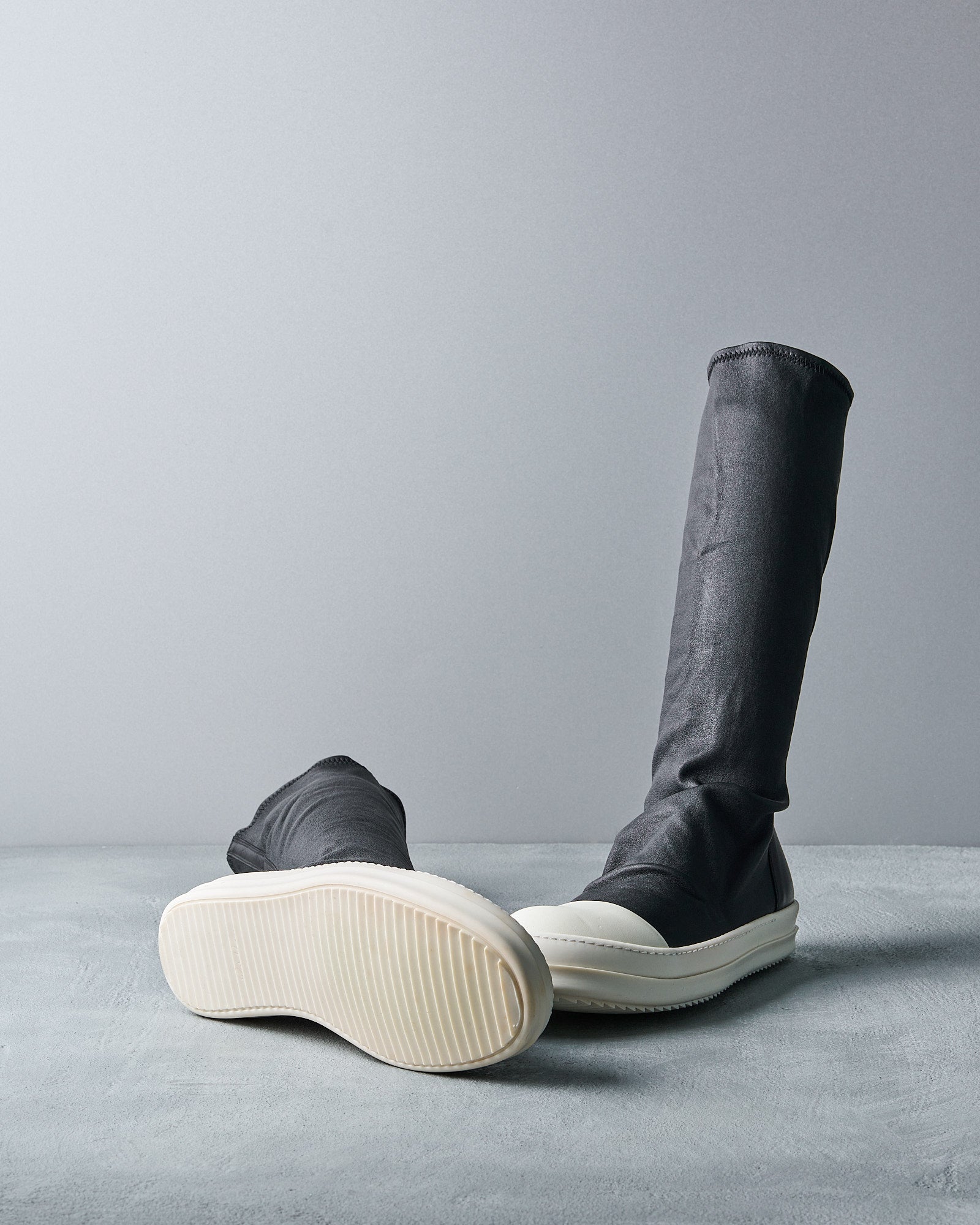 Black high-top Rick Owens sneakers with white soles, one lying down and the other upright against a gray backdrop, convey a modern, sleek style.