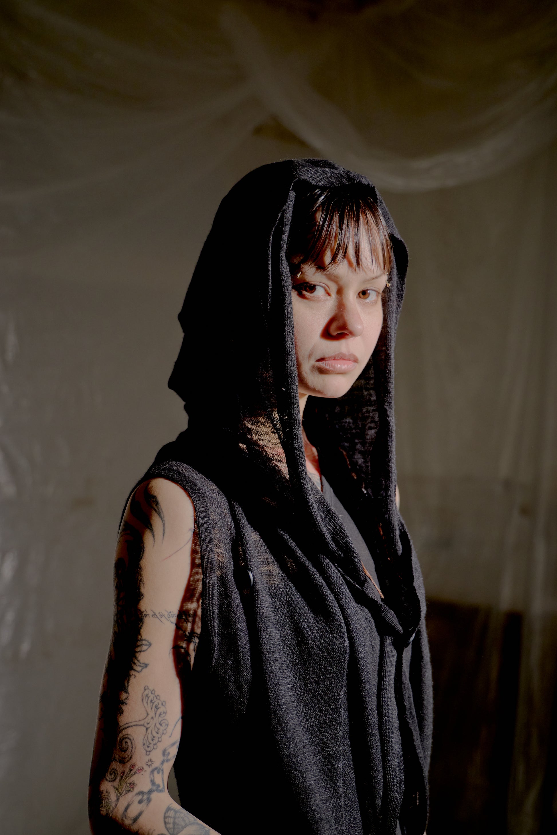 Person wearing a dark hooded Rick Owens garment with tattoos on arms against a plain background