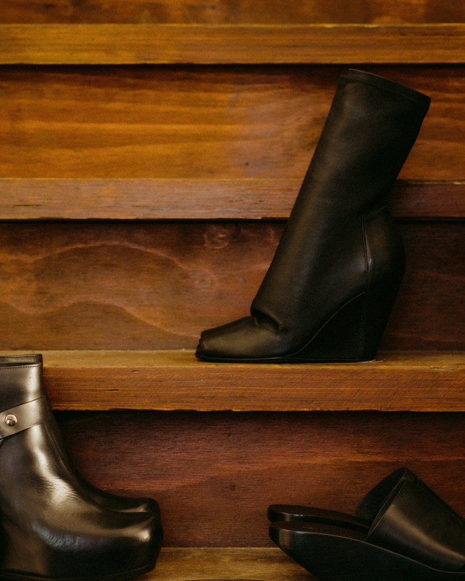 A stylish black Rick Owens wedge boot is displayed on wooden shelves, suggesting a modern and elegant fashion choice against a warm, rustic backdrop.