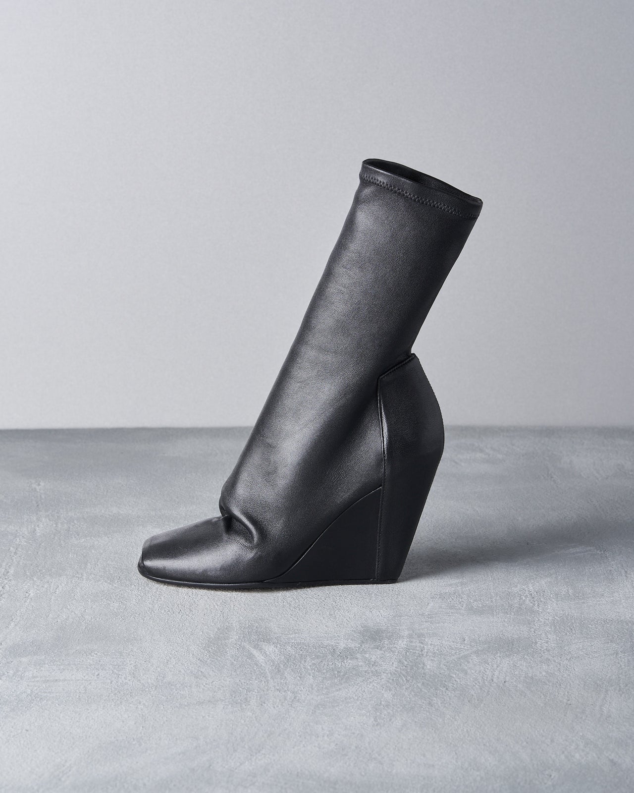 A stylish black Rick Owens wedge boot is displayed on wooden shelves, suggesting a modern and elegant fashion choice against a warm, rustic backdrop.