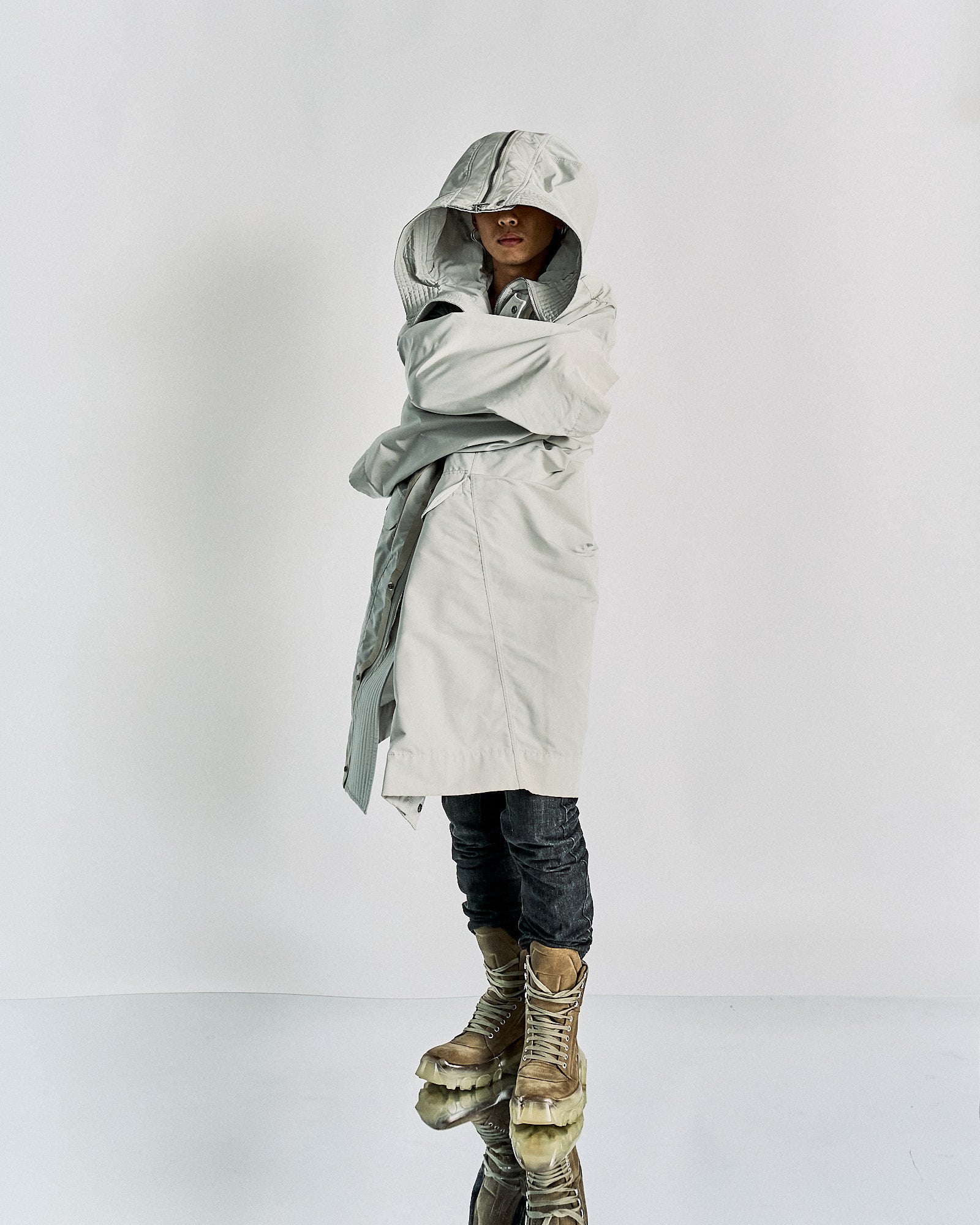 Person in a gray hooded Rick Owens coat stands on a reflective surface, arms crossed. Wears dark pants and tan boots. Mysterious and edgy atmosphere.