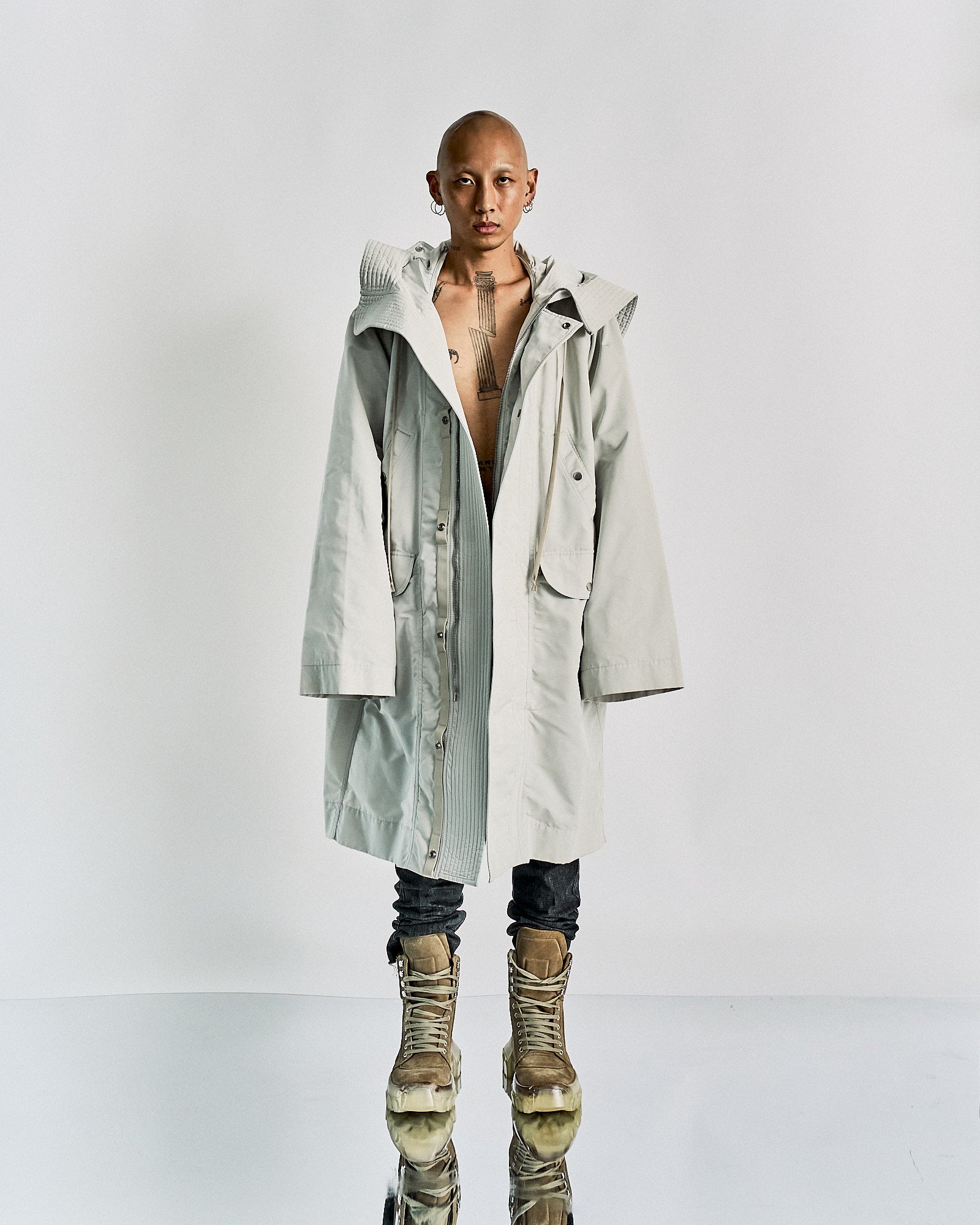 Person in a gray hooded Rick Owens coat stands on a reflective surface, arms crossed. Wears dark pants and tan boots. 