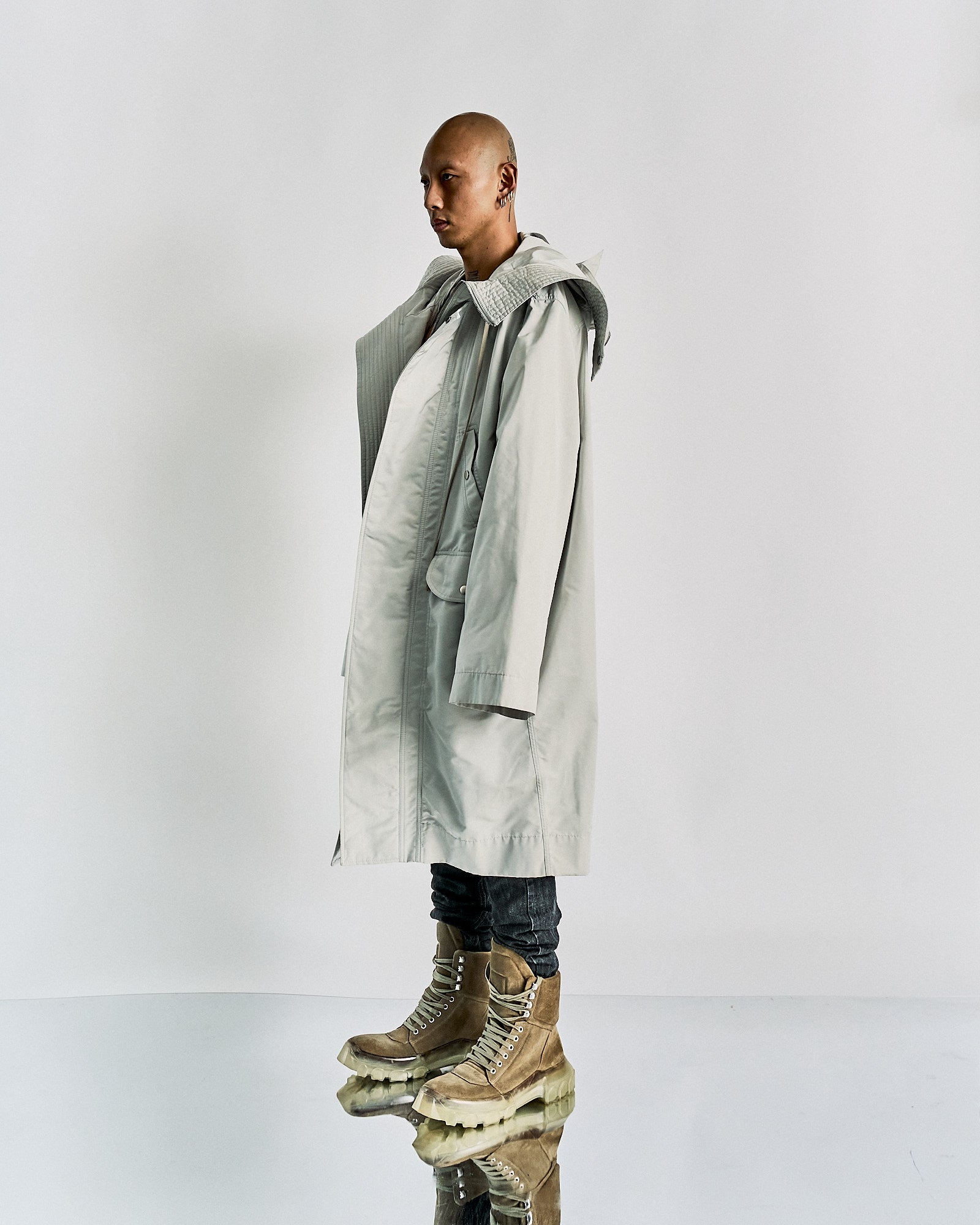 A person stands sideways wearing an oversized light grey Rick Owens coat with a textured collar, paired with slim dark pants and chunky beige boots.