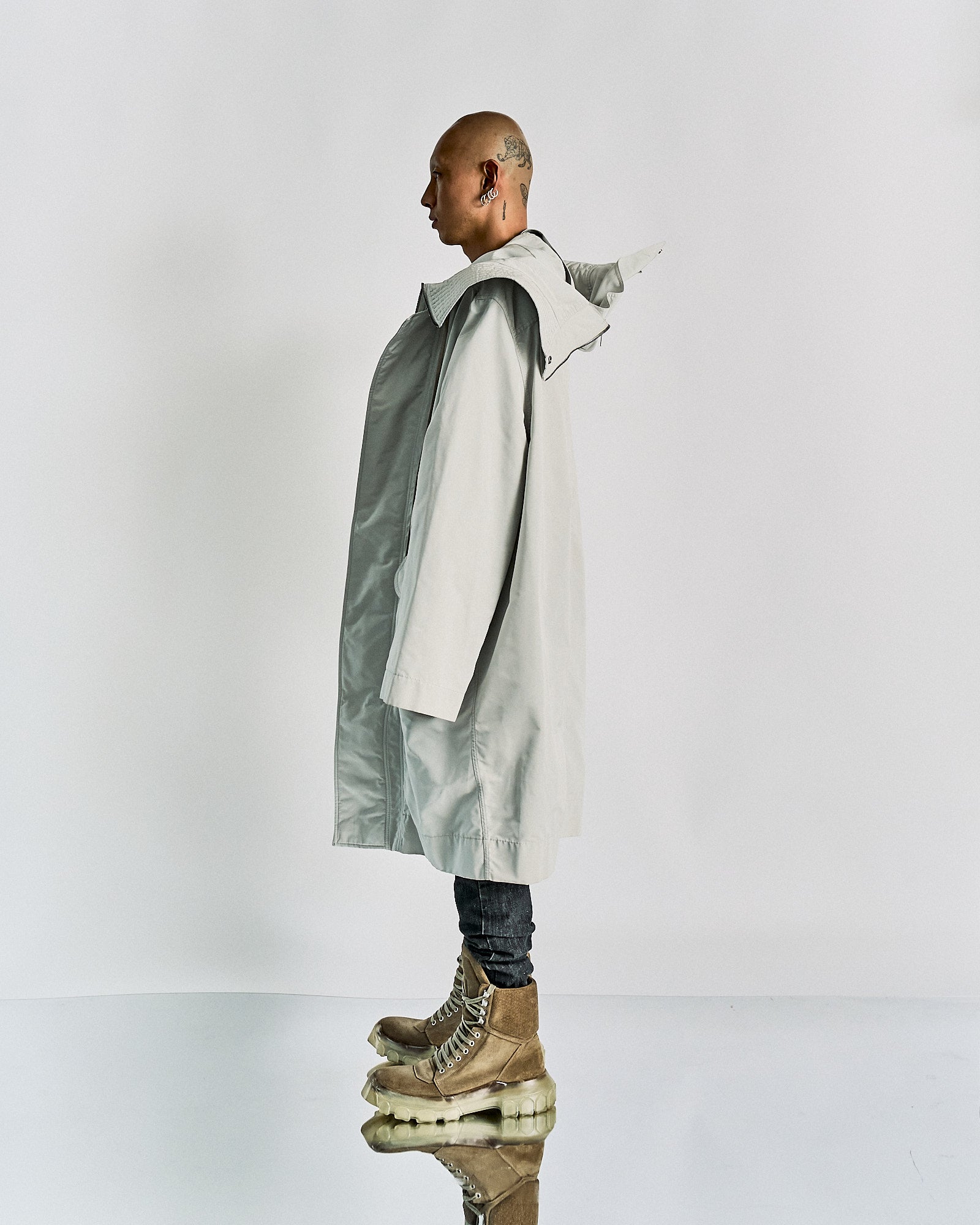 A person stands sideways wearing an oversized light grey Rick Owens coat with a textured collar, paired with slim dark pants and chunky beige boots. The tone is modern and edgy.