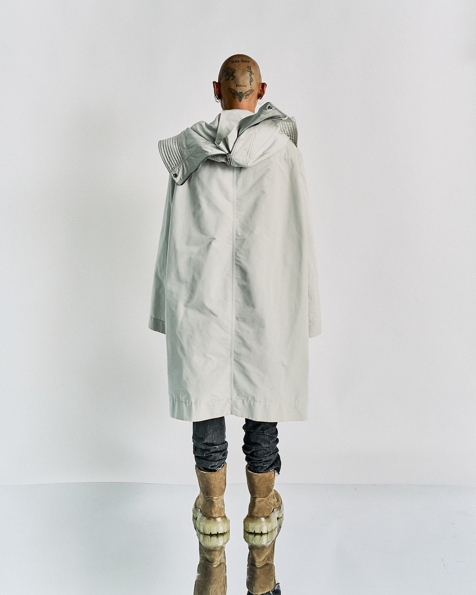 A person stands with their back to the camera, wearing a long white Rick Owens coat and futuristic boots. The head is shaved, showing tattoos.