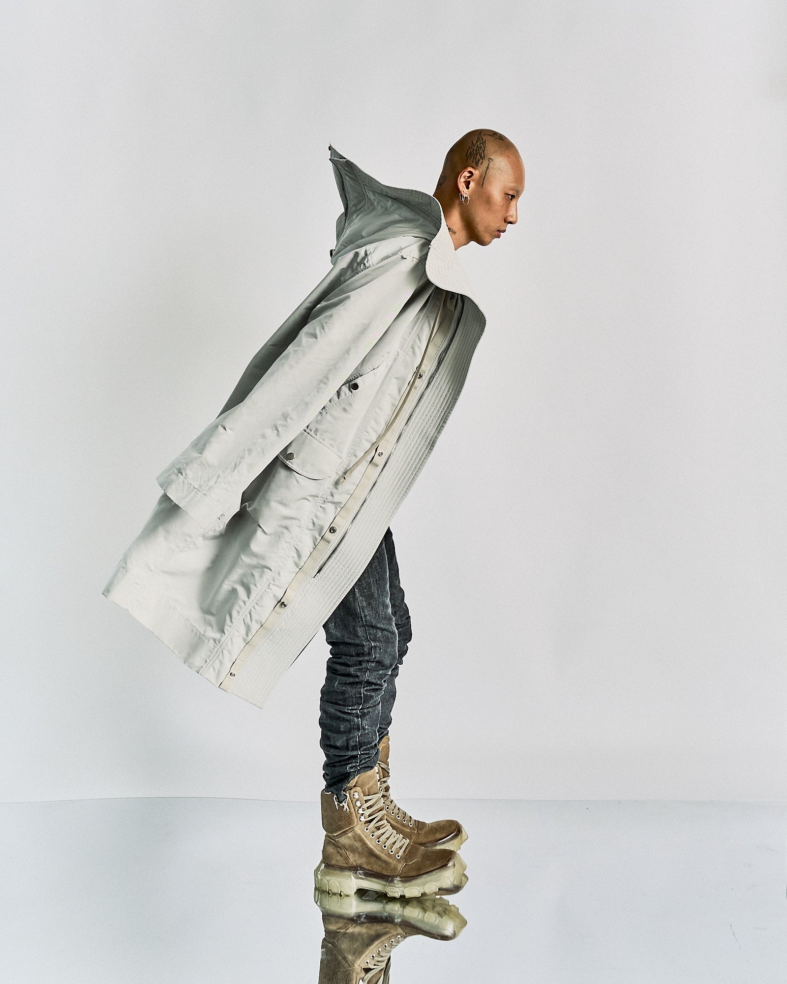 A person dressed in an oversized light grey Rick Owens coat and worn jeans stands hunched forward on translucent platform boots, creating a futuristic and surreal atmosphere.