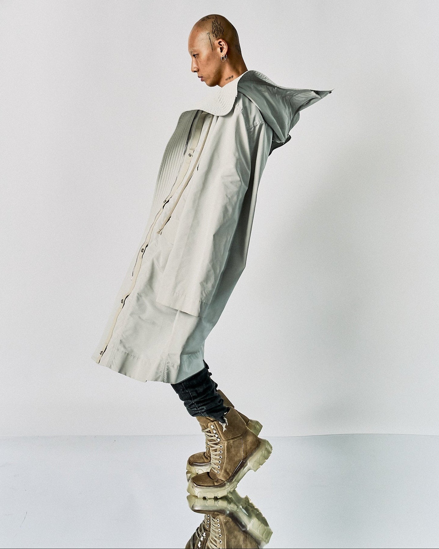 A person in a wide, light grey coat leans dramatically forward on a reflective surface. They wear beige boots, exuding a bold, avant-garde fashion vibe.