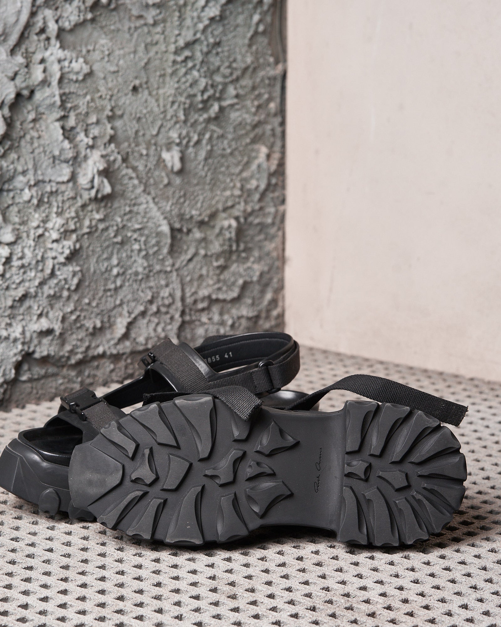 Black Rick Owens sandals with rugged, textured soles are placed on a patterned surface. A rustic, weathered wall forms the backdrop, adding a contrasting element.