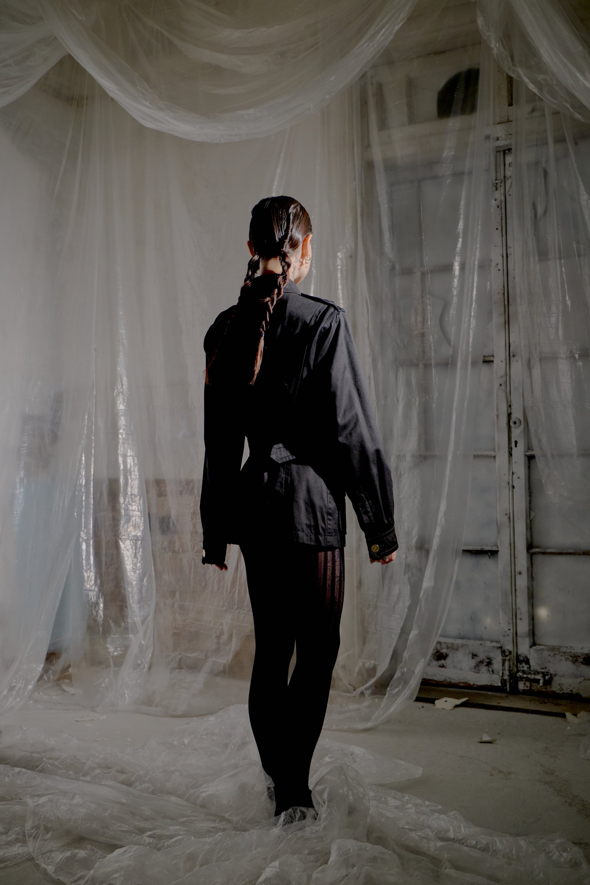 Person standing in a room with sheer white curtains and a rustic door wearing a black Saint Laurent outfit.