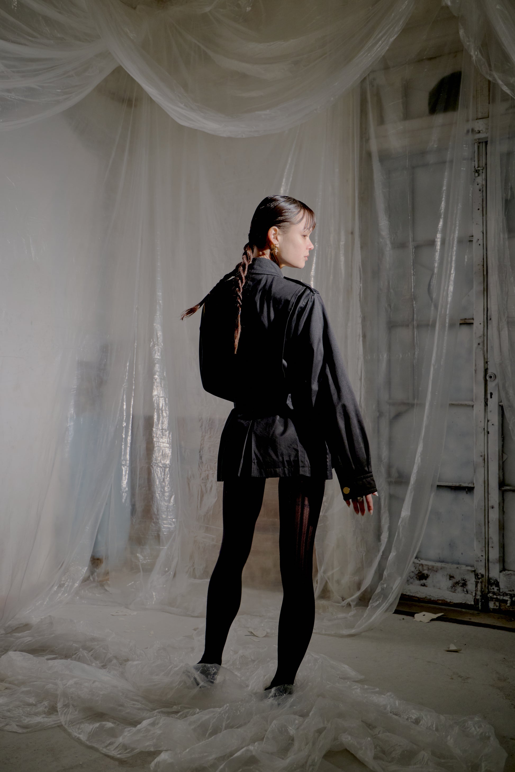 Person standing in a room with sheer white curtains and a rustic door wearing a dark Saint Laurent jacket. 
