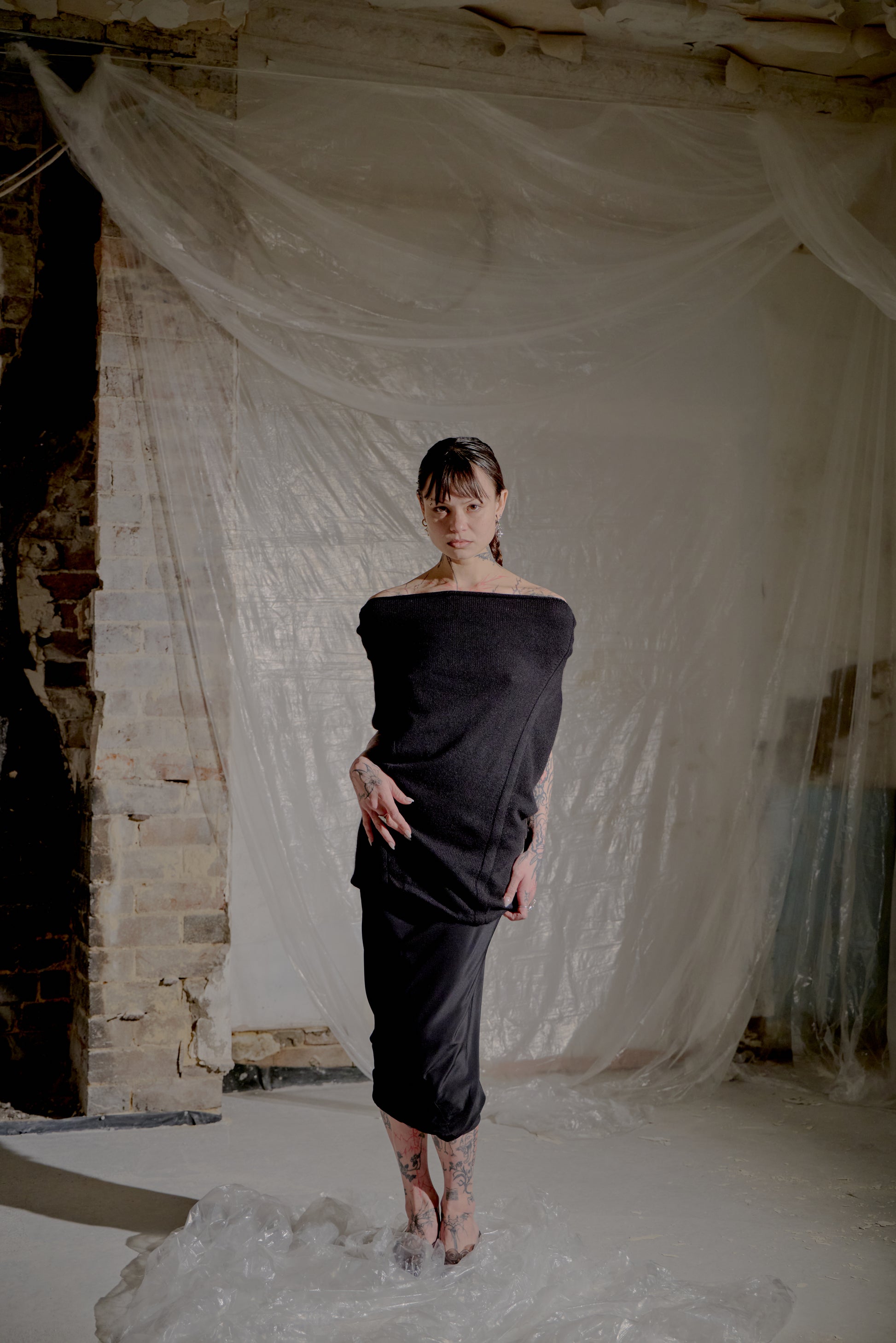Woman in a black off-shoulder Saint Laurent dress standing in a room with white fabric and brick walls.