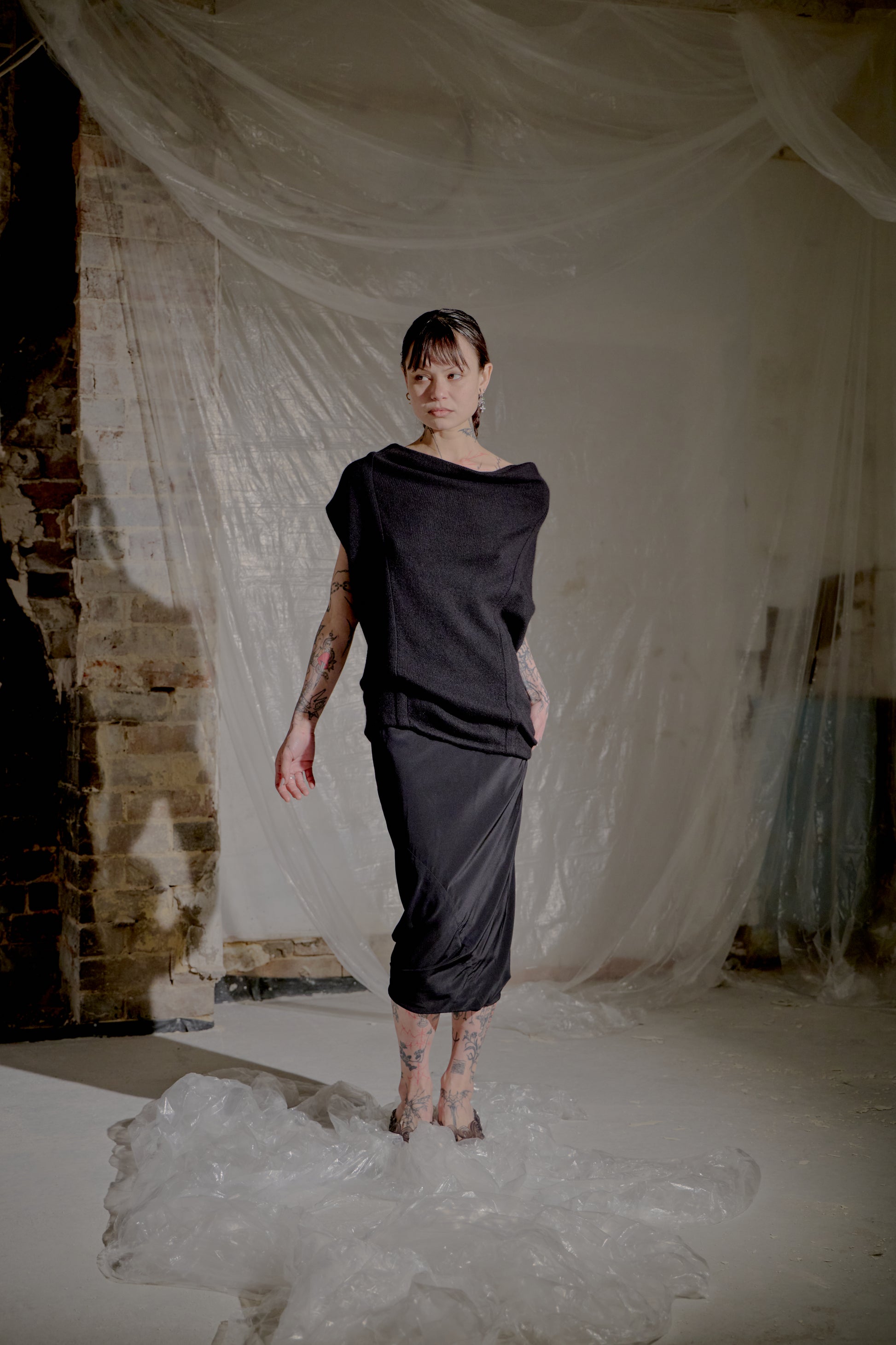 Person wearing a black Saint Laurent outfit standing in a room with white fabric and brick wall.