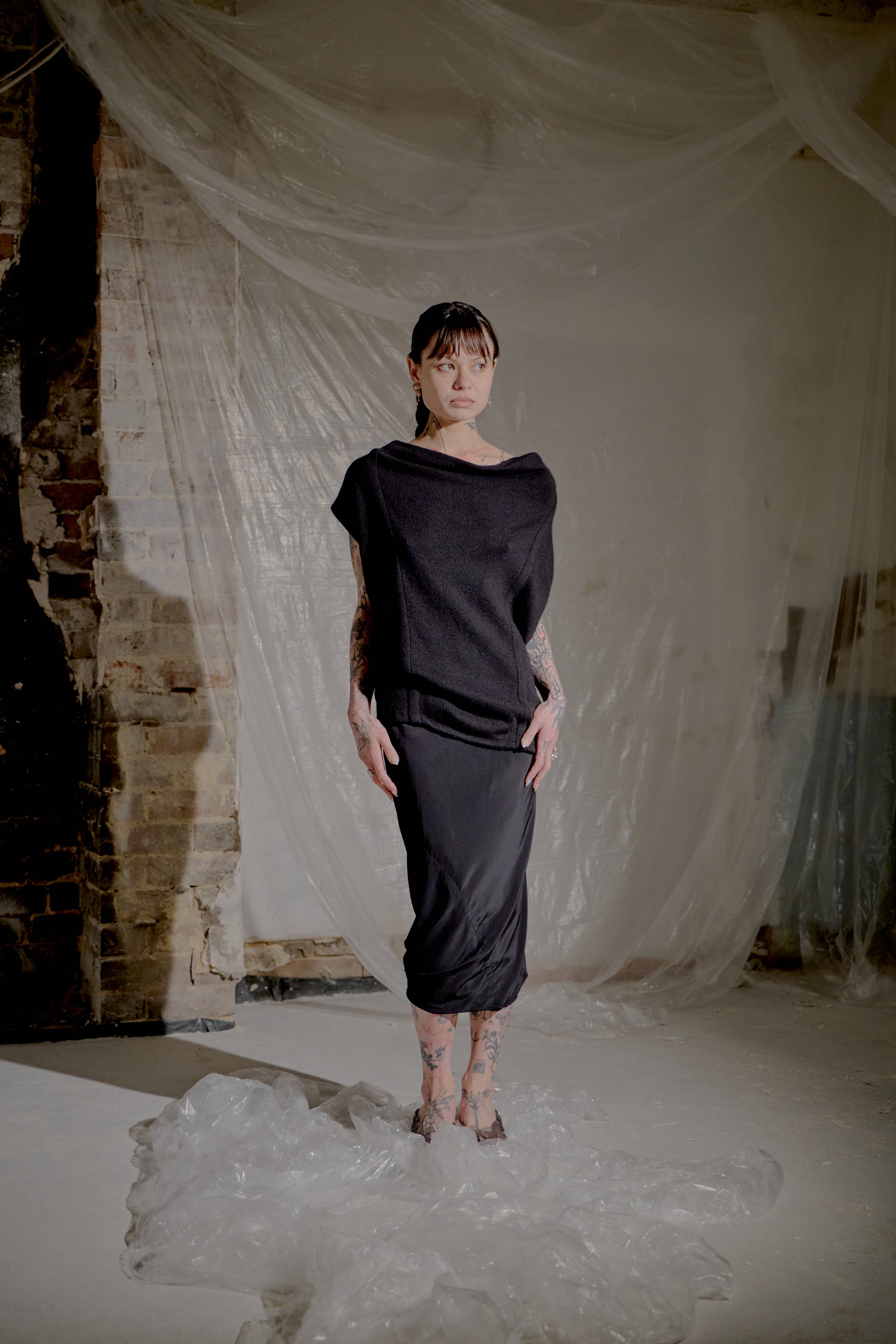 Person wearing a black Saint Laurent outfit standing in a room with white fabric and brick wall.