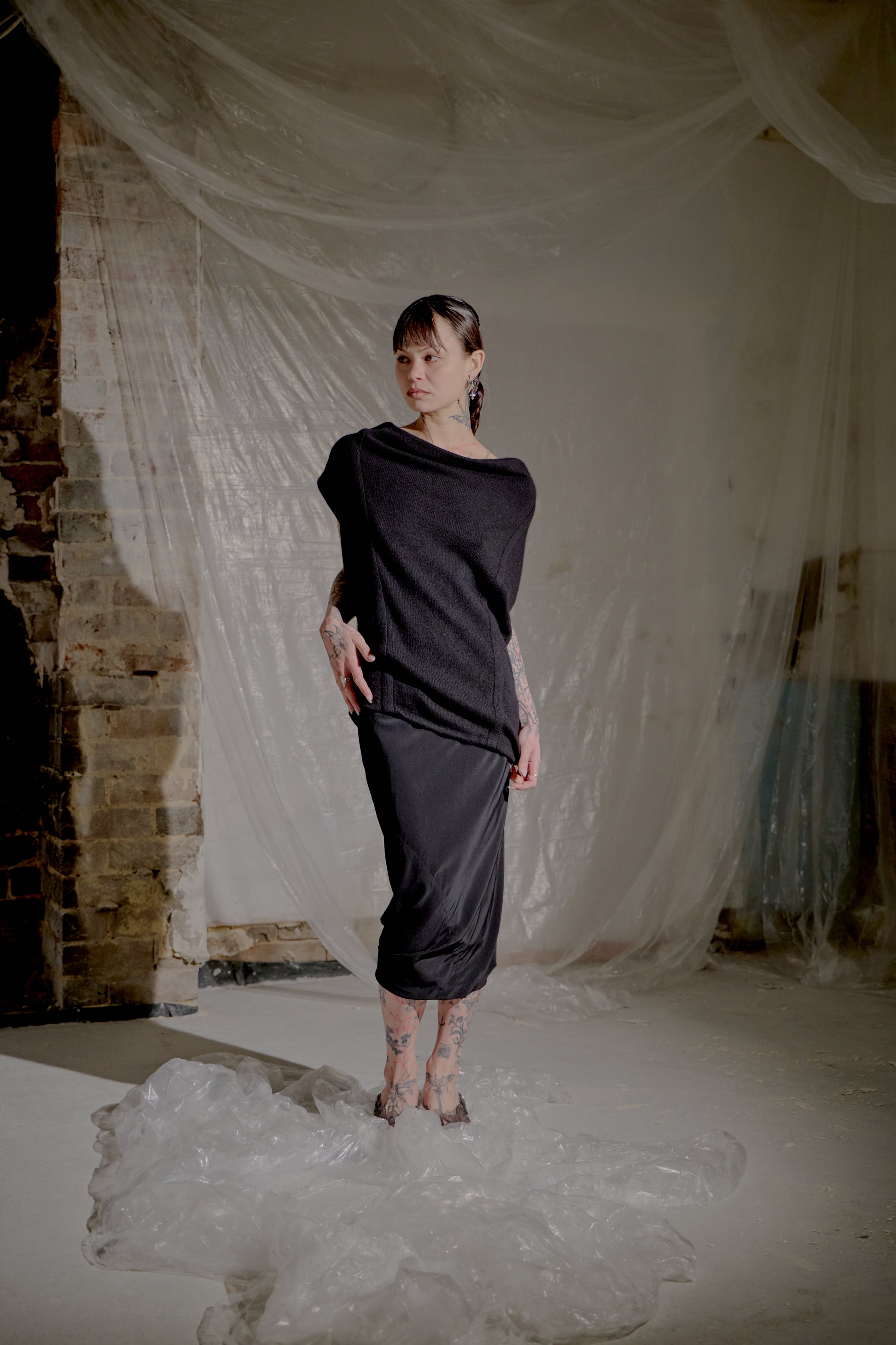 Woman in a black Saint Laurent outfit standing in a room with white fabric and brick wall.