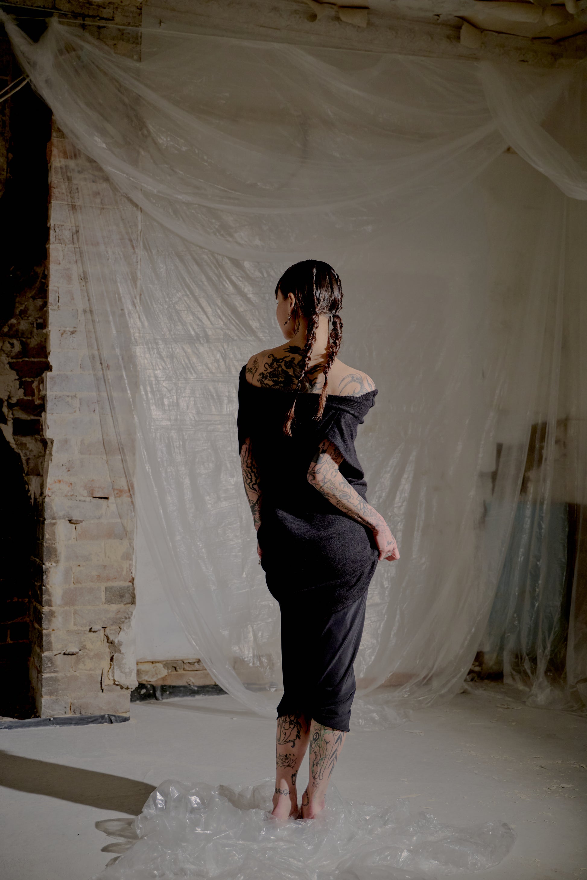 Person in a black Saint Laurent dress standing in a room with white fabric and brick wall.