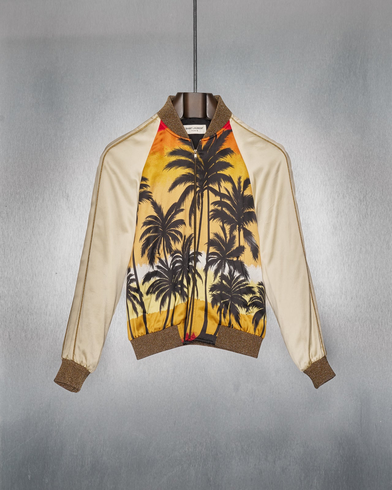 A model walks the Saint Laurent runway wearing white sunglasses, an ornate gold jacket with palm tree designs, a textured scarf, and black leather pants with patches. 
