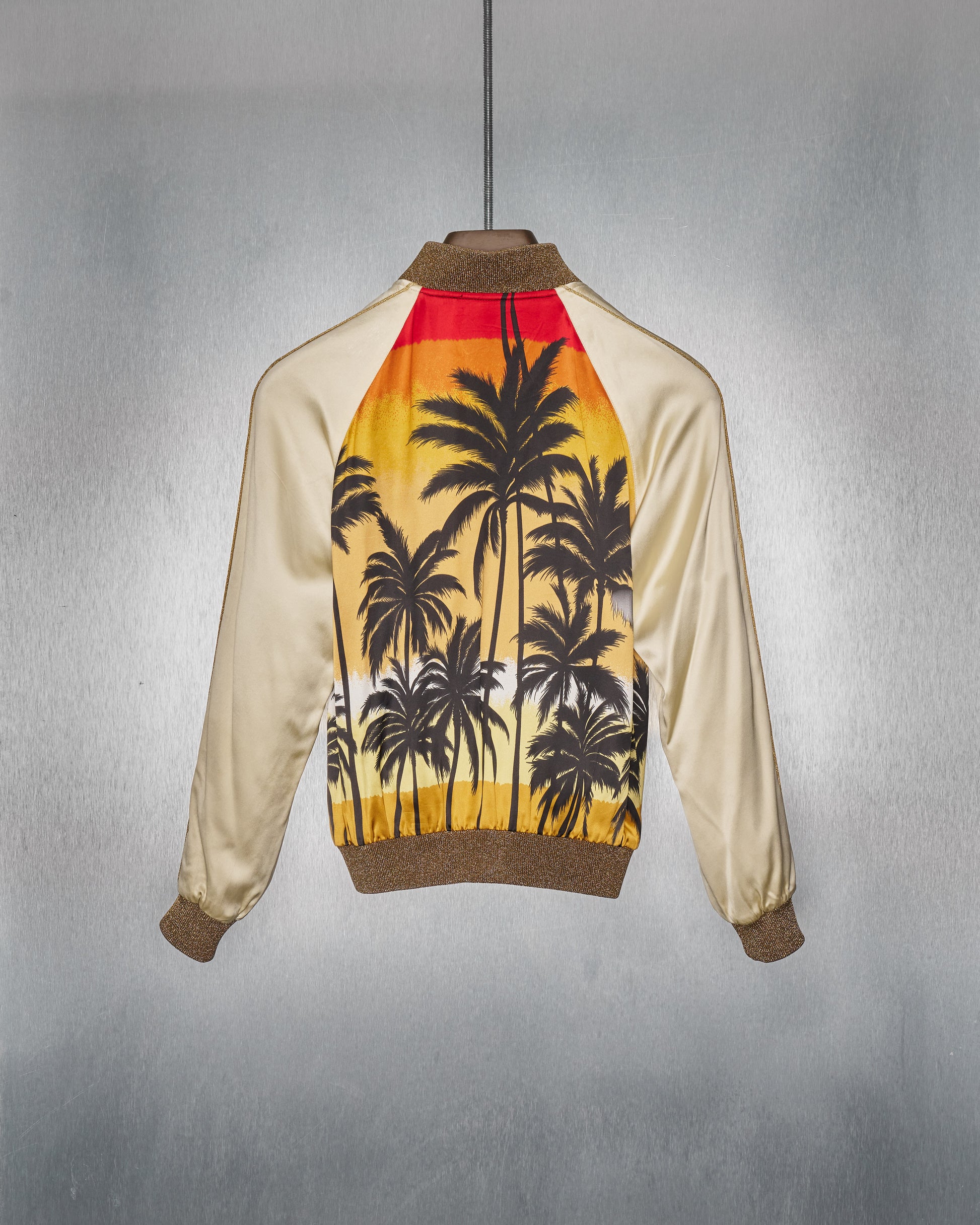 A satin Saint Laurent bomber jacket is displayed against a silver background. Its back features a vibrant sunset design with palm silhouettes, evoking a tropical feel.