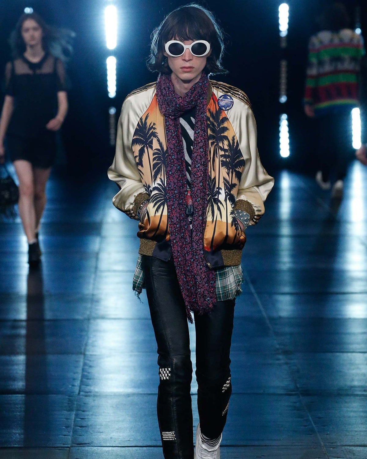 A model walks the Saint Laurent runway wearing white sunglasses, an ornate gold jacket with palm tree designs, a textured scarf, and black leather pants with patches. 