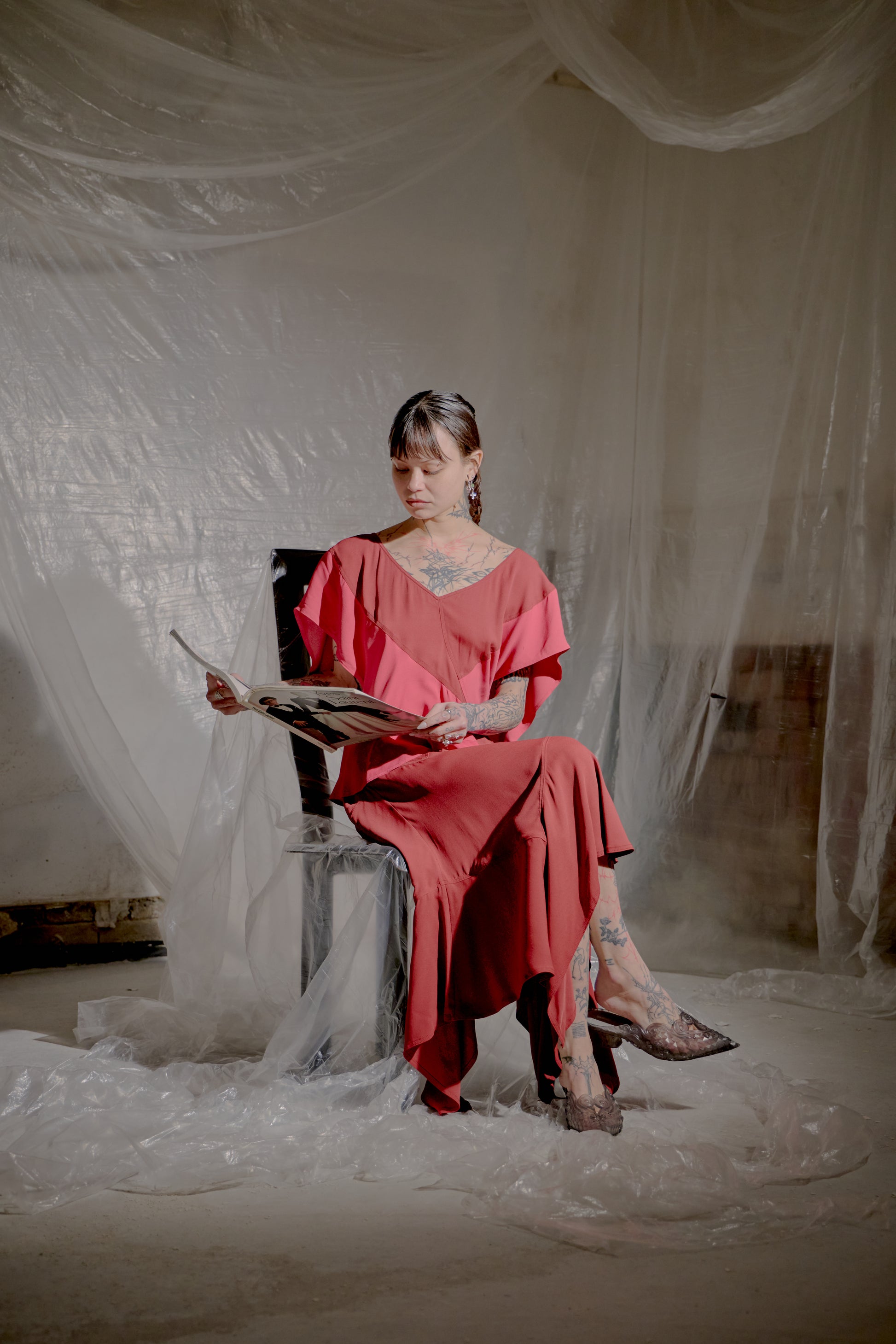 Woman in a red Saint Laurent dress sitting on a chair holding a book in a room with white draped fabric.