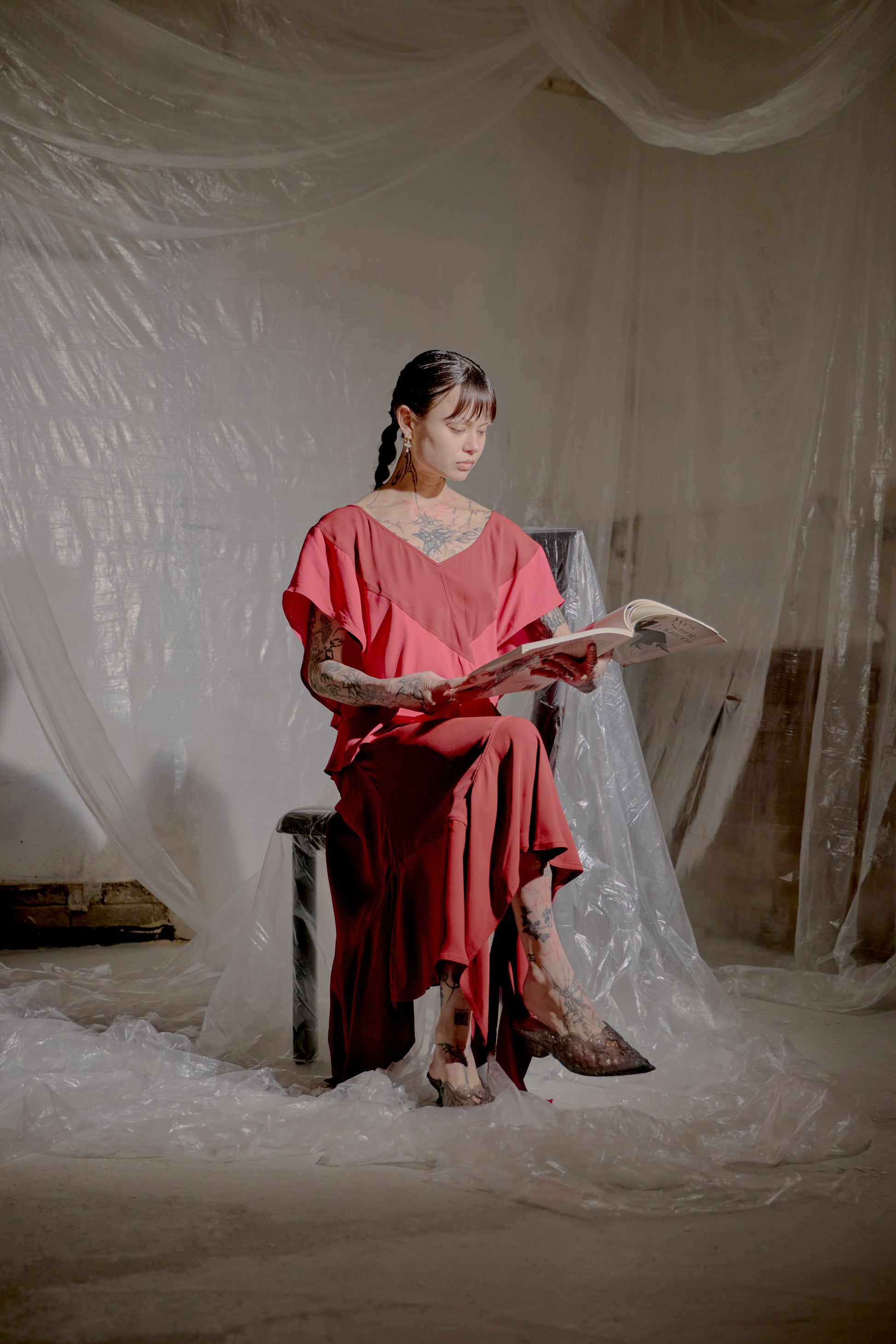 Woman in a red Saint Laurent dress sitting on a chair reading a book against a white draped background