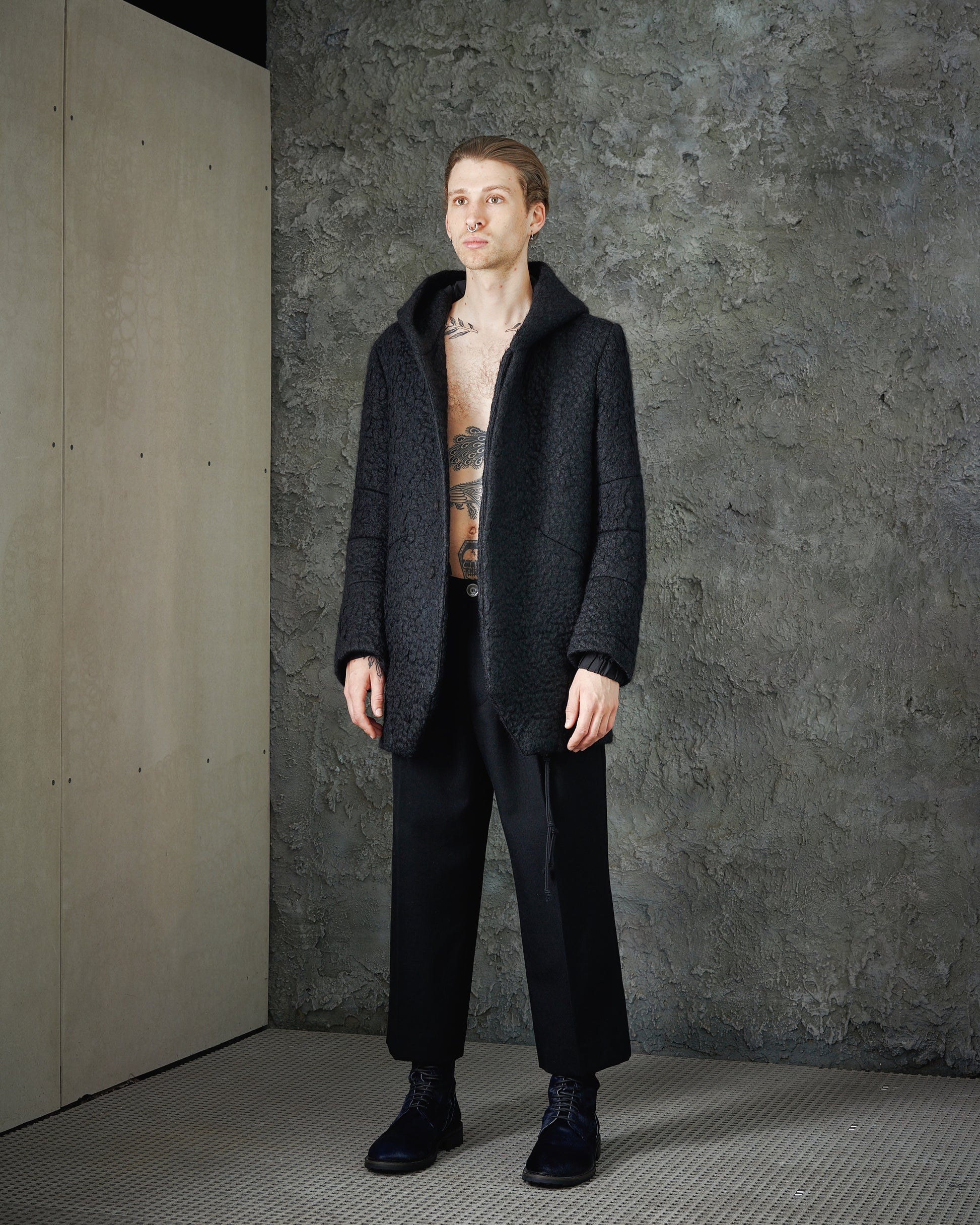 A person wearing a black hooded Song For The Mute mohair cocoon crinkle coat with elasticized cuffs and two hip pockets, standing in a grey, textured background.
