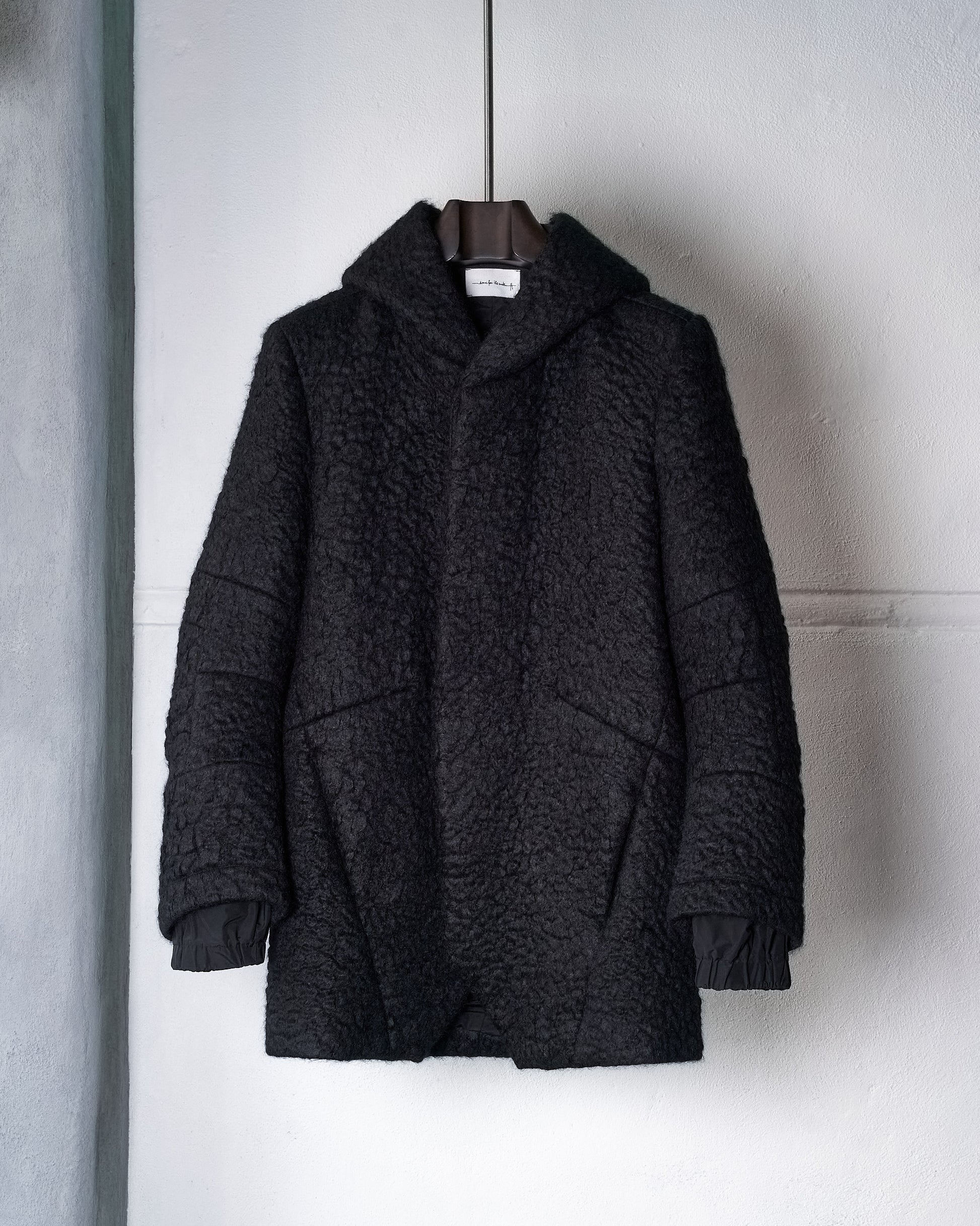 Black textured Song For The Mute coat with a hood, hanging on a dark hanger against a white wall. The cozy fabric conveys warmth and style.