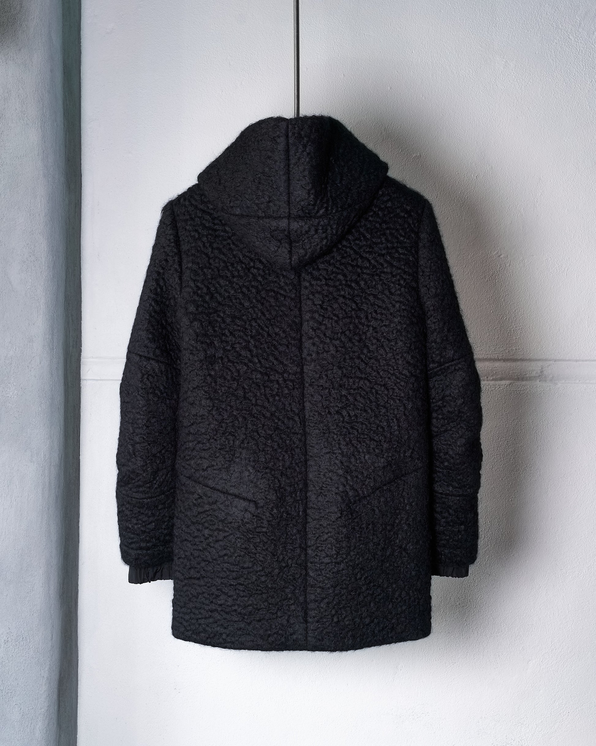 Reverse of a black textured Song For The Mute coat with a hood, hanging on a dark hanger against a white wall. The cozy fabric conveys warmth and style.