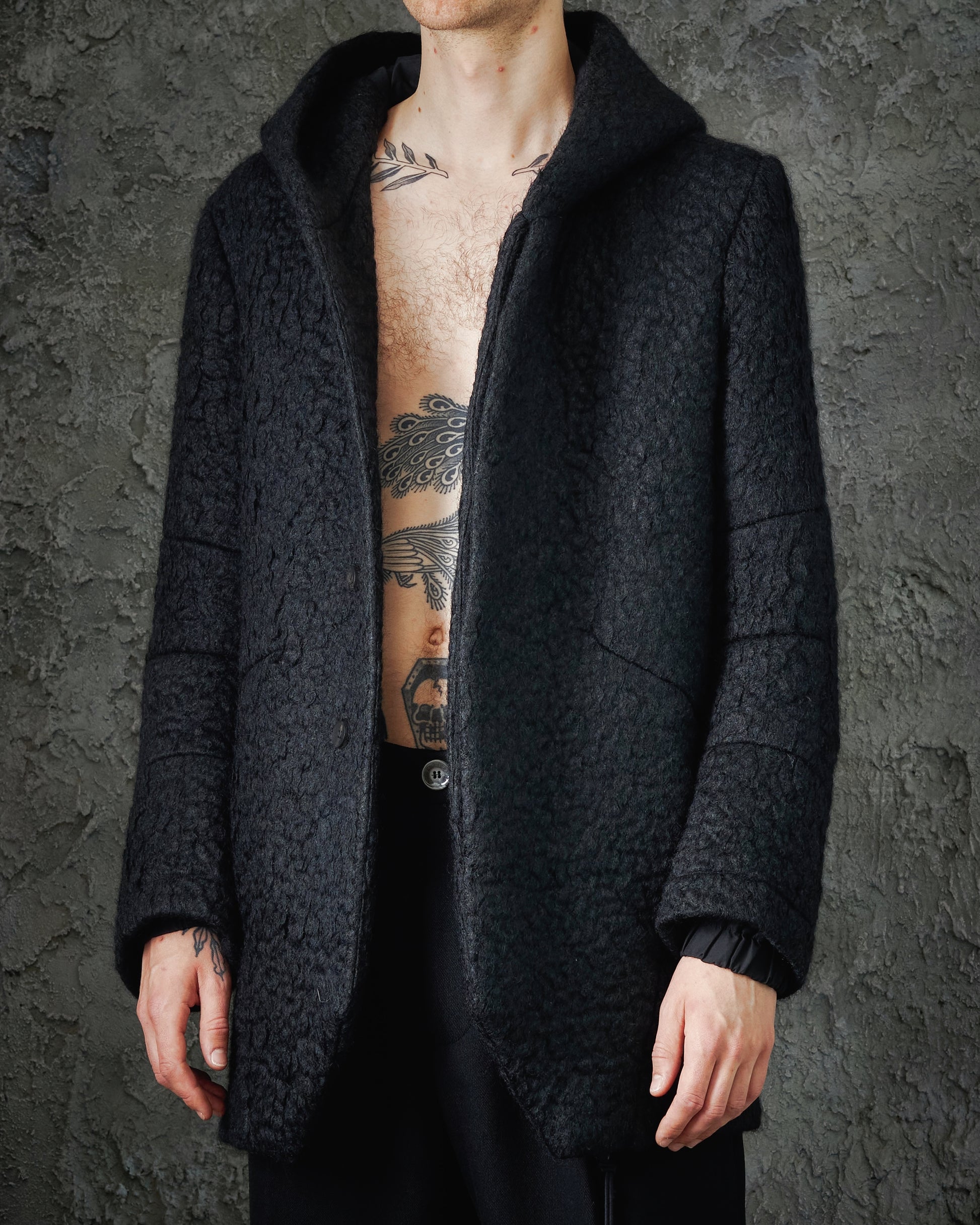 Person in a textured black Song For The Mute coat with a hood stands against a rough, gray wall. Their chest tattoos are partially visible, creating a bold, edgy tone.