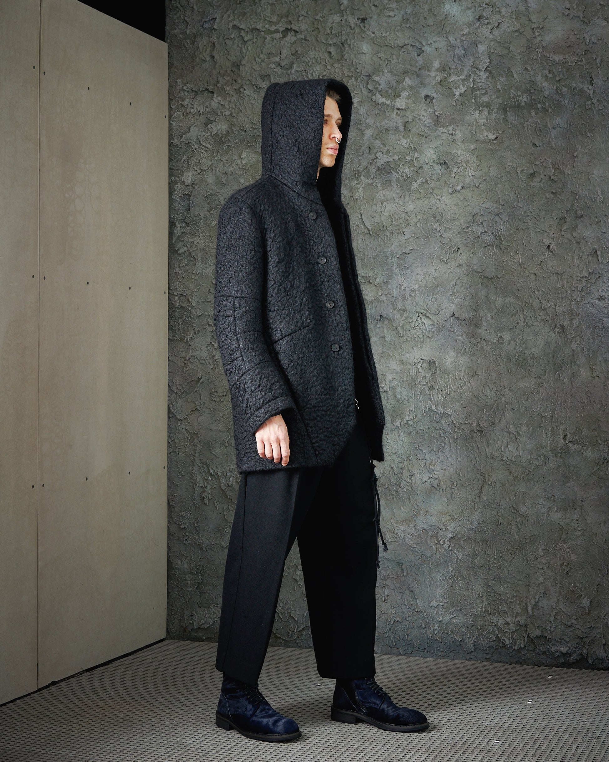 A person stands in profile, wearing a textured dark Song For The Mute coat with a hood, black pants, and shoes. The backdrop is a rough gray wall, creating a moody, urban vibe.