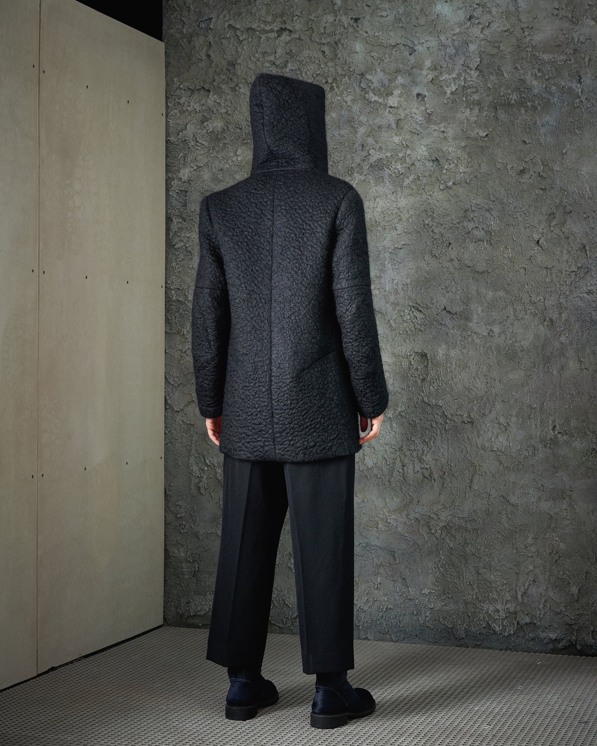 Person in a textured dark Song For The Mute coat with hood, black pants, and shoes stands against a rough gray wall. The setting has a somber, minimalist tone.