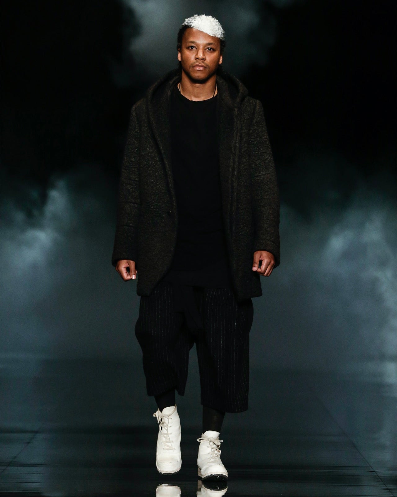 Lupe Fiasco wearing a black Song For The Mute outfit with white shoes on a dark background