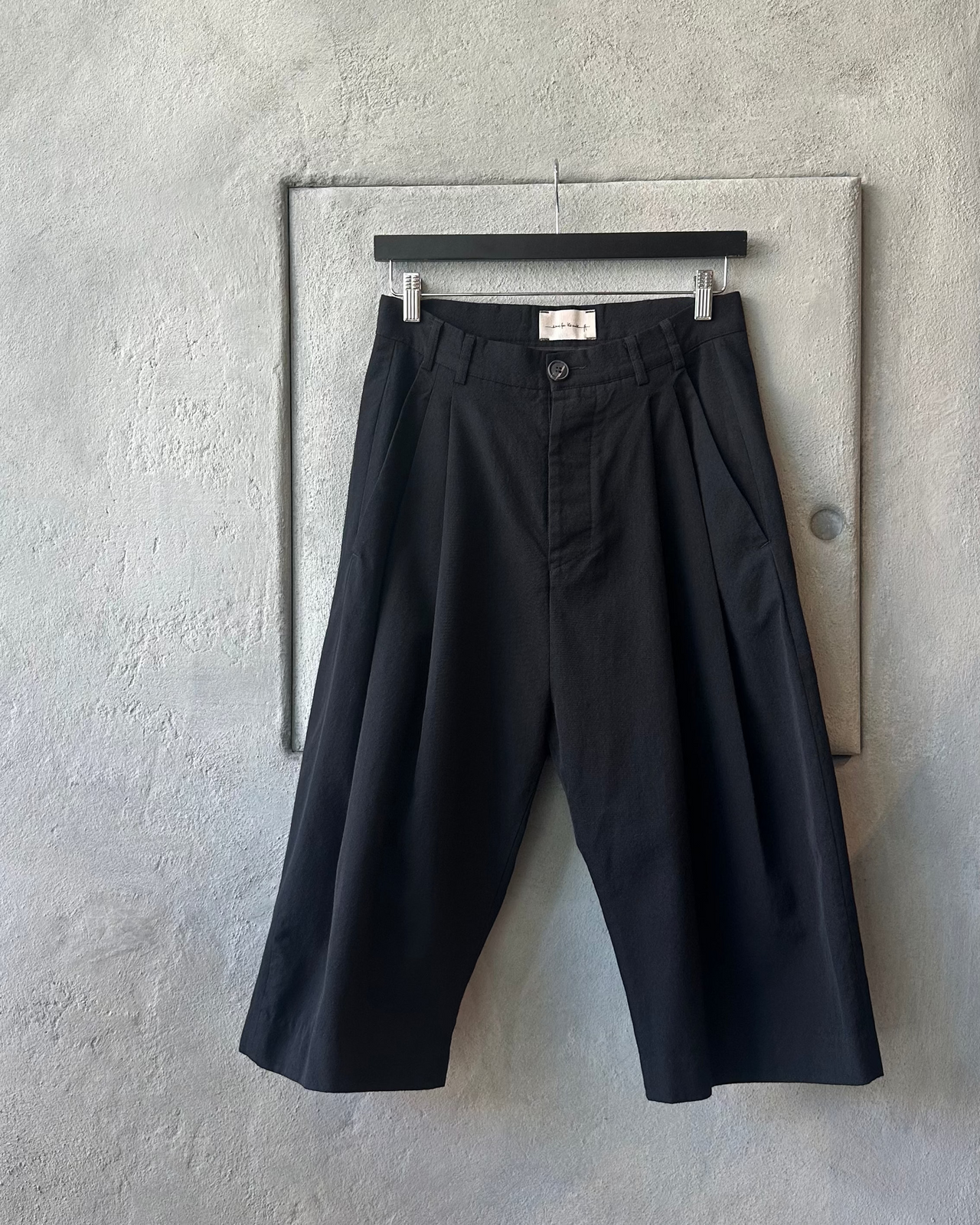 Black Song For The Mute pants hanging on a hanger against a gray wall