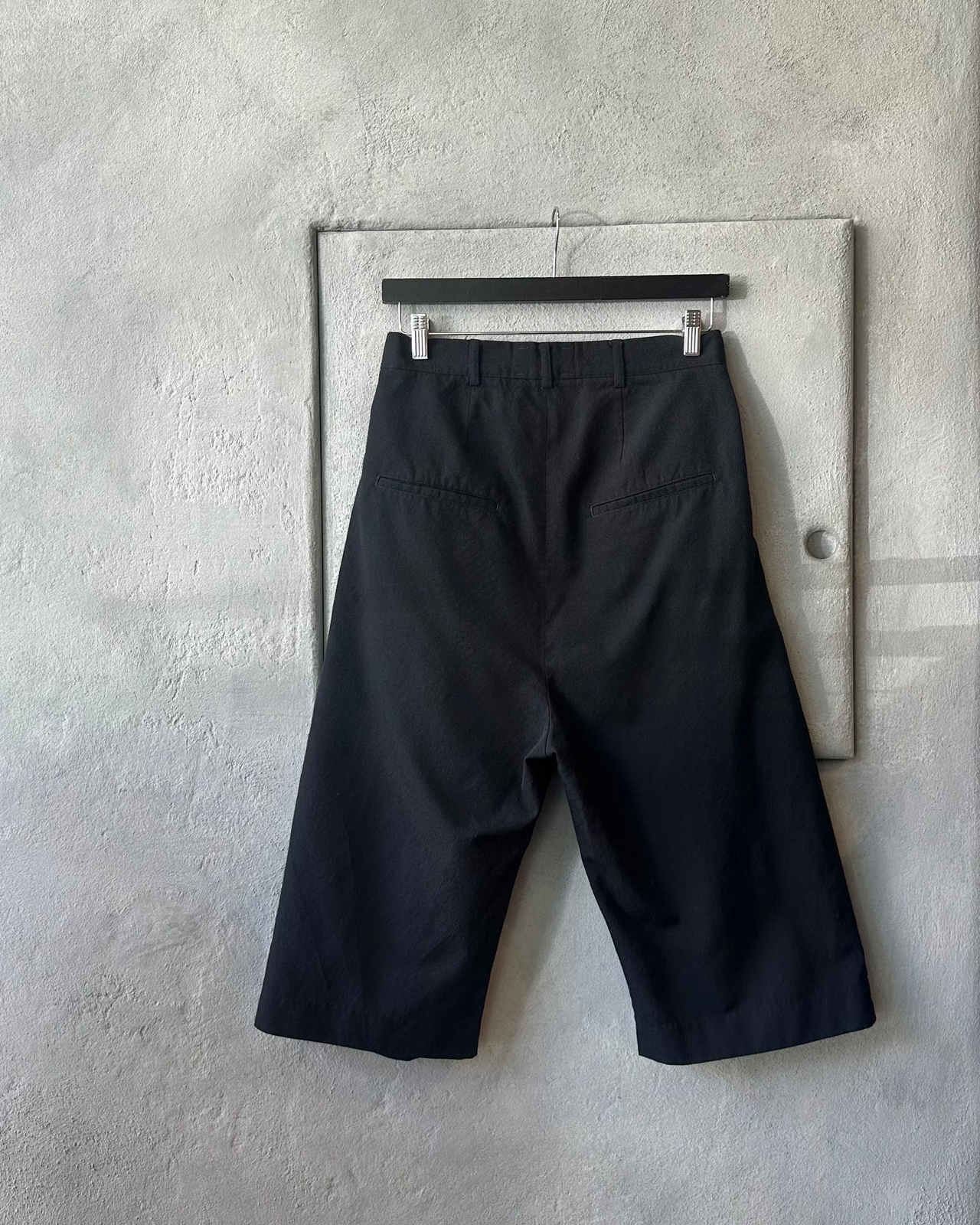Black Song For The Mute pants hanging on a hanger against a gray wall