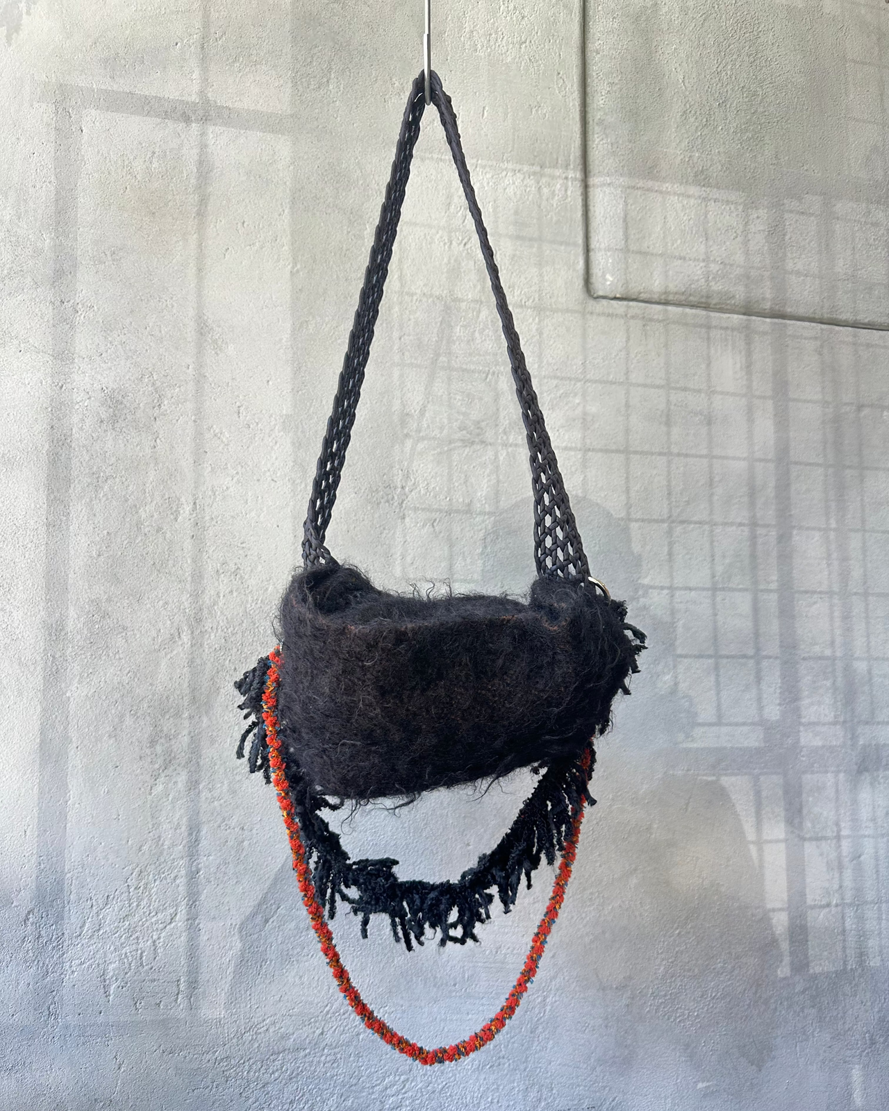 Black woven Song For The Mute bag with red rope hanging against a textured wall.