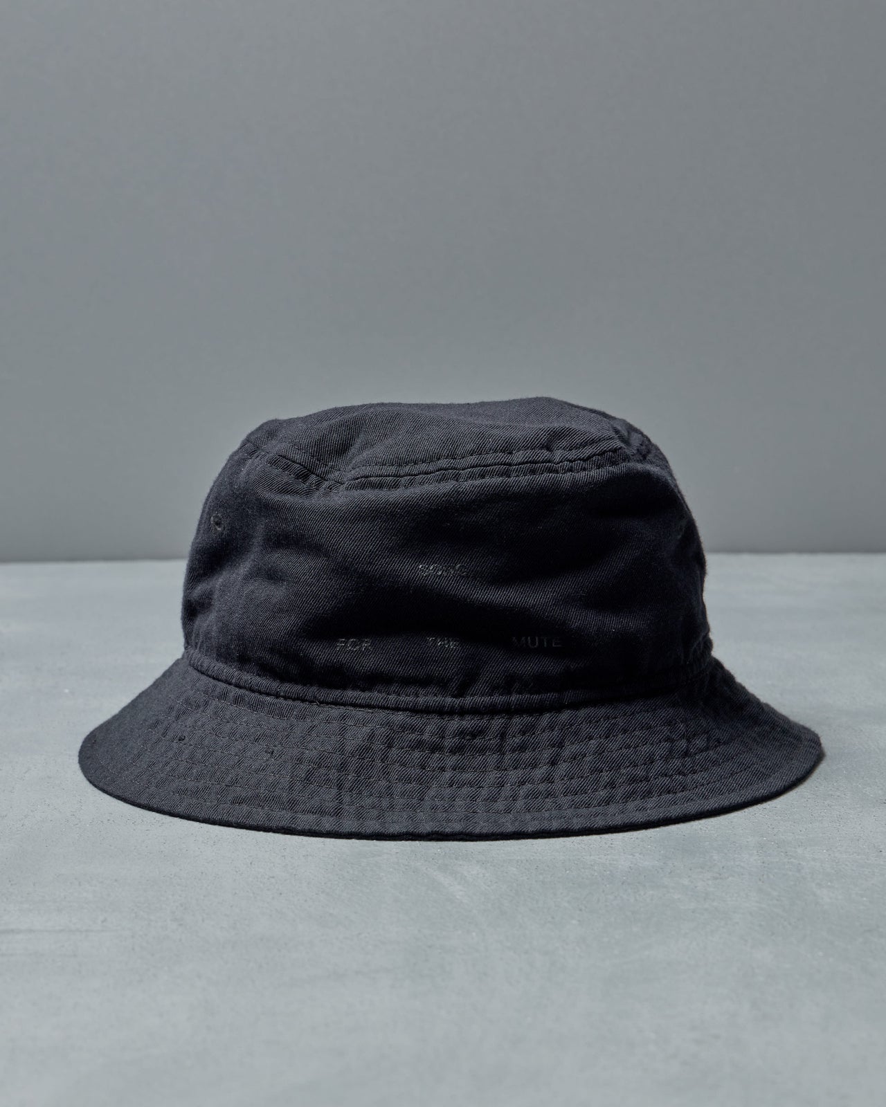 A black Song For The Mute x New Era bucket hat with subtle embroidered details sits on a light gray surface. 