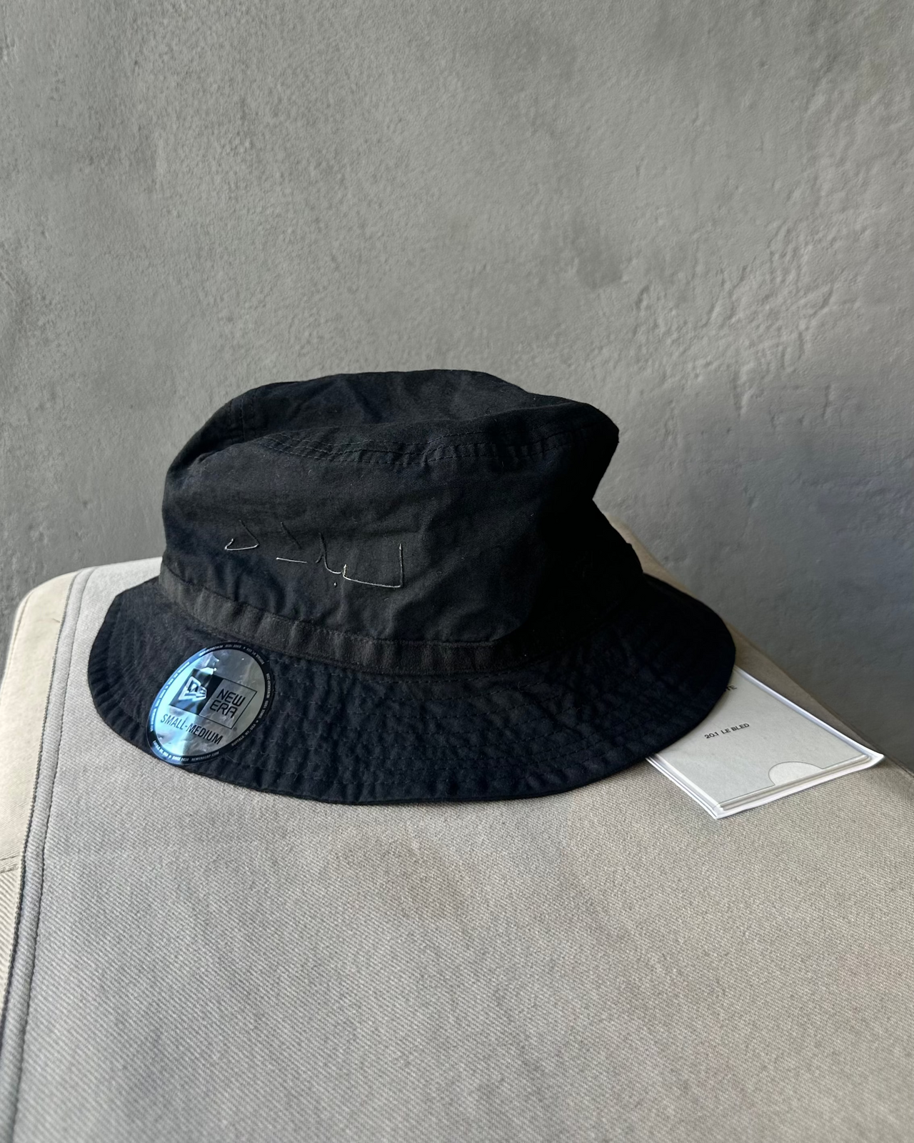 Black Song For The Mute x New Era bucket hat hanging on a metal rod against a light gray wall.