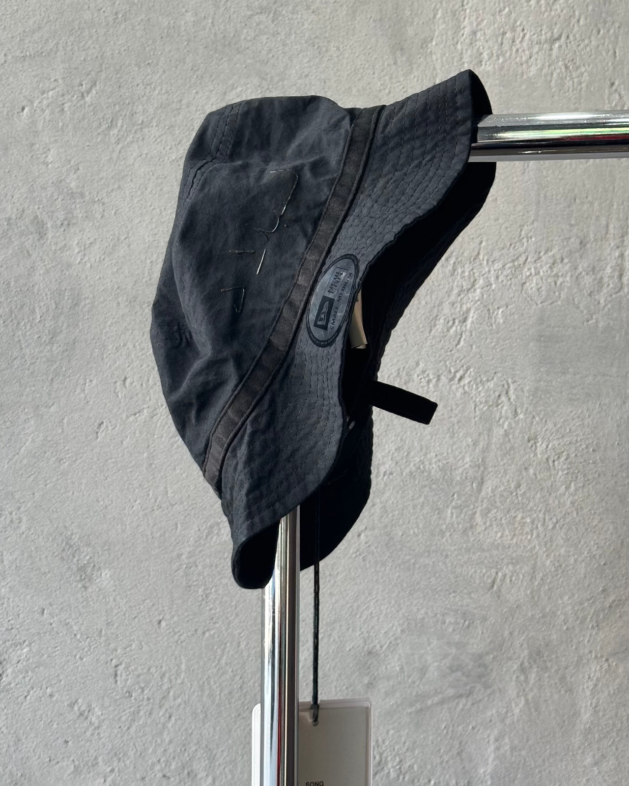 Black Song For The Mute x New Era bucket hat hanging on a metal rod against a light gray wall.