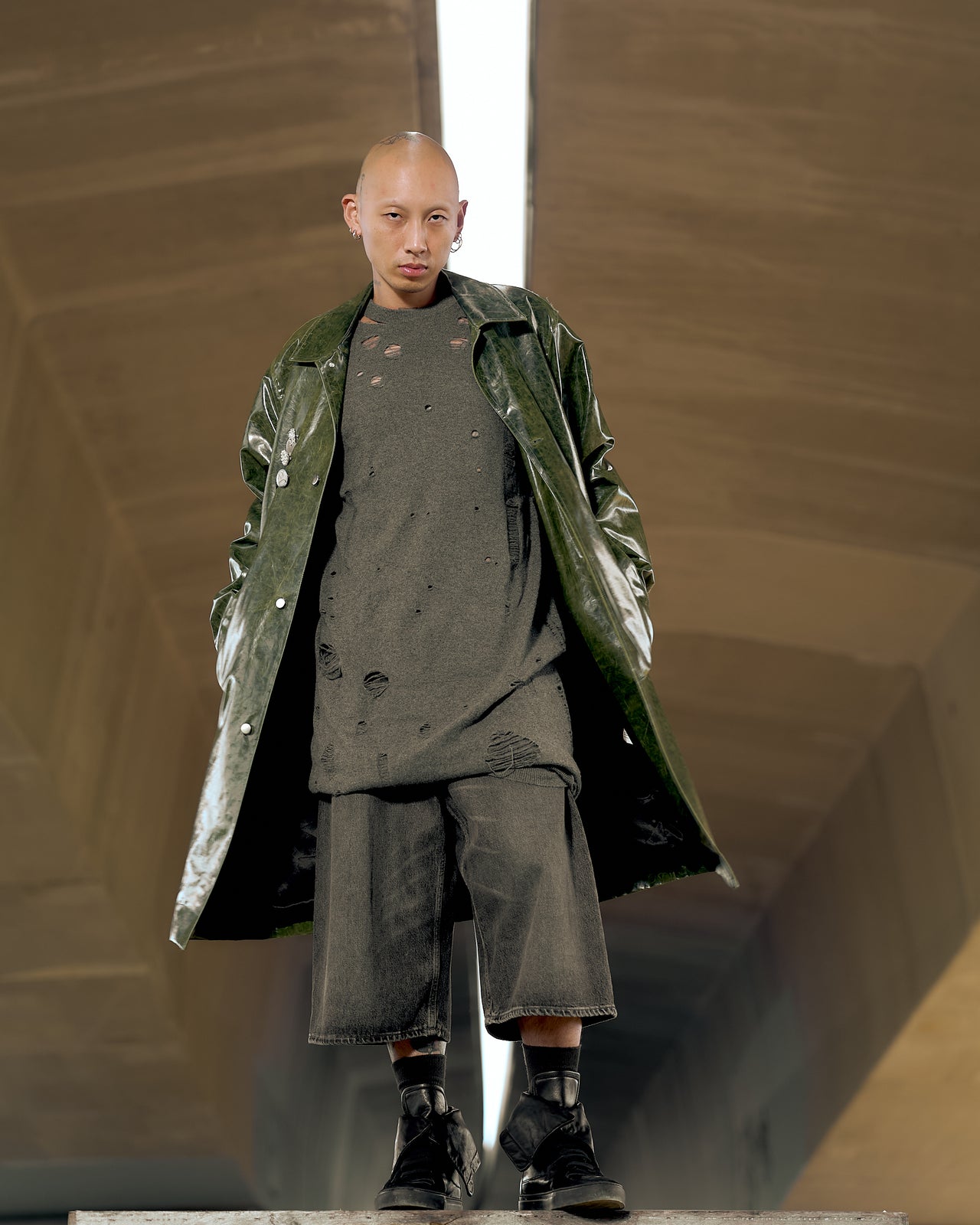 A model wearing a knee-length Song For The Mute car coat in green, with metal buttons and brooches, standing in a reflective space.