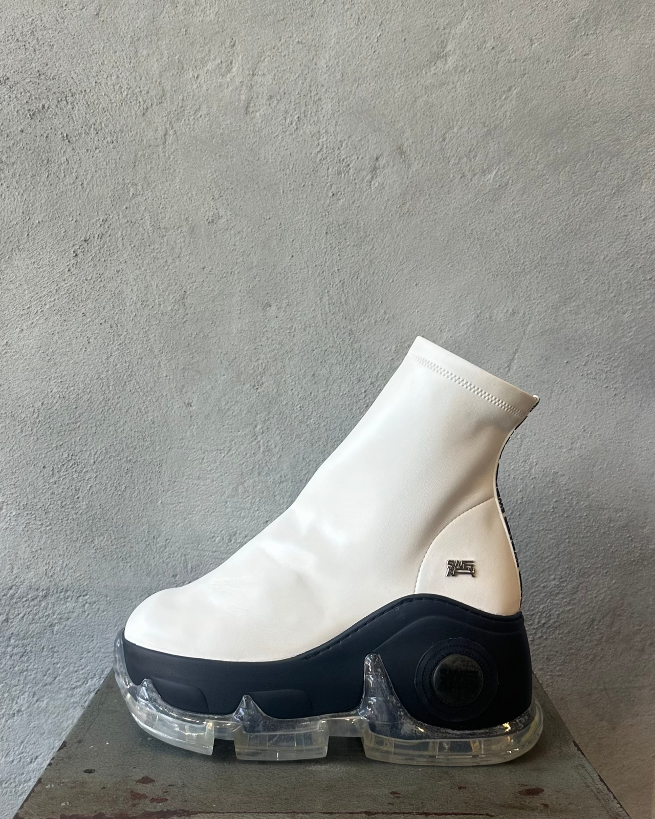 White and black high-top Swear London platform sneaker on a metal stool against a gray concrete wall.