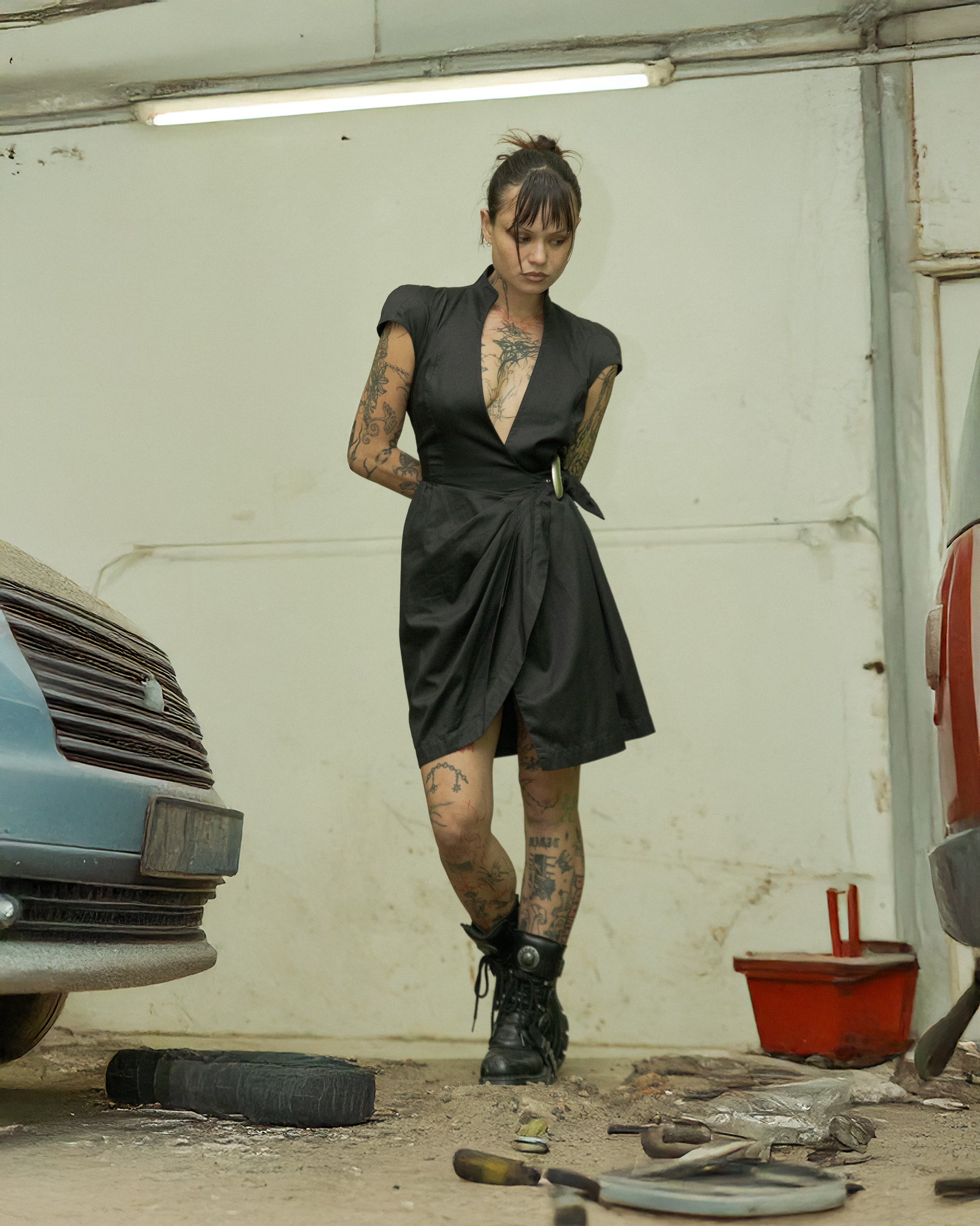 A person standing in a garage wearing a black belted Thierry Mugler dress with cap sleeves, paired with black boots.