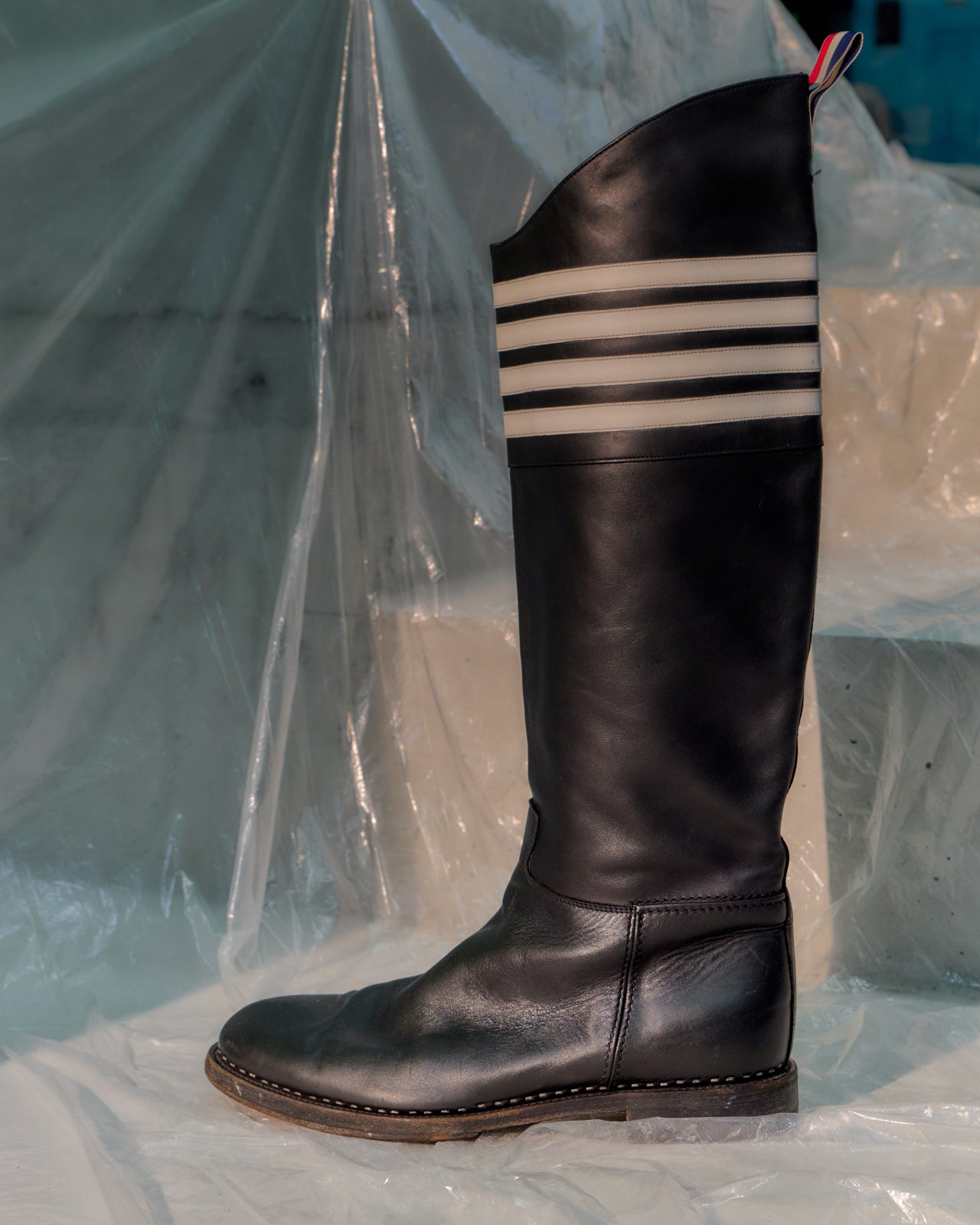 Black knee-high Thom Browne boot with three white stripes on a light blue background