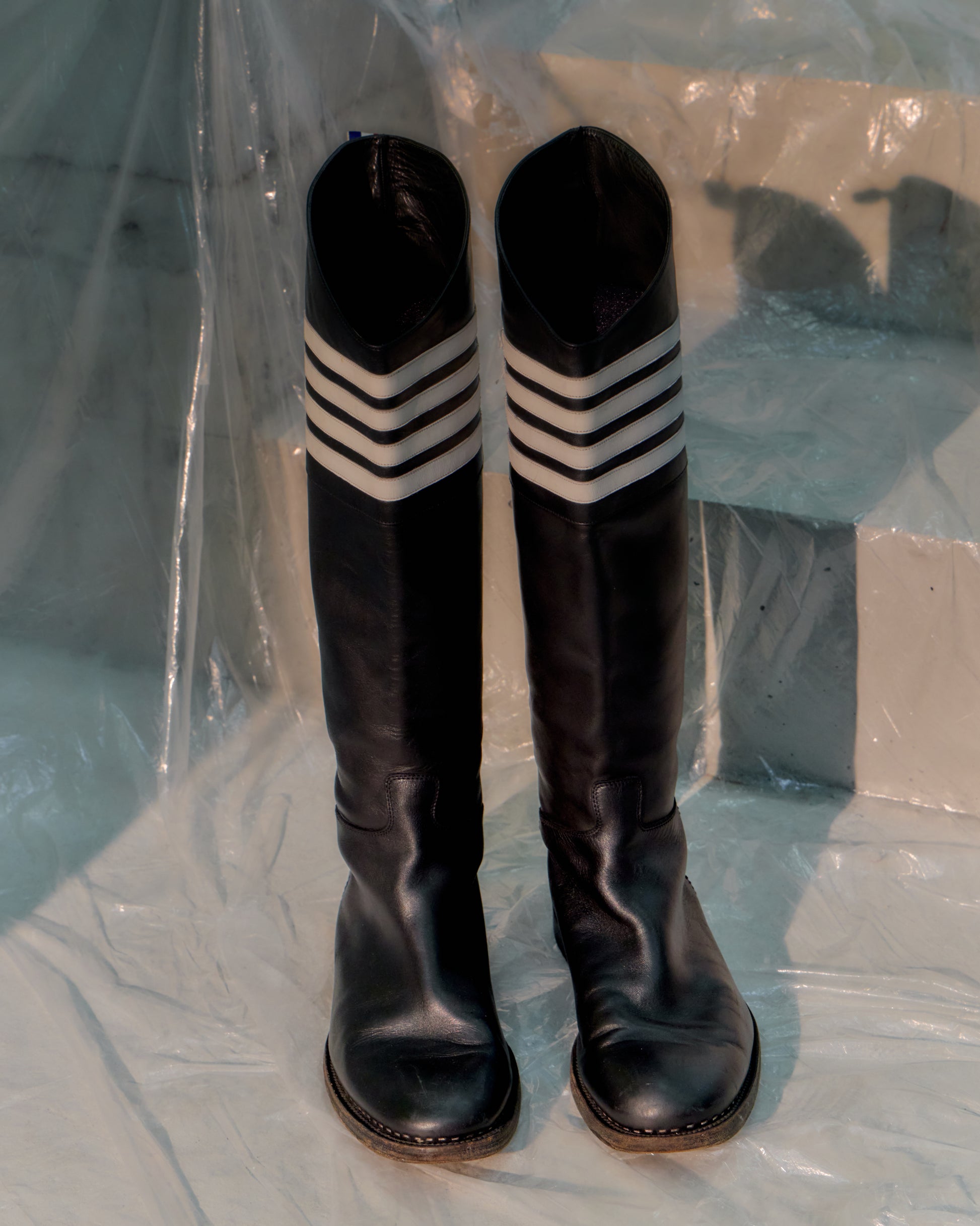 Black knee-high Thom Browne boots with white striped socks on a clear plastic sheet.