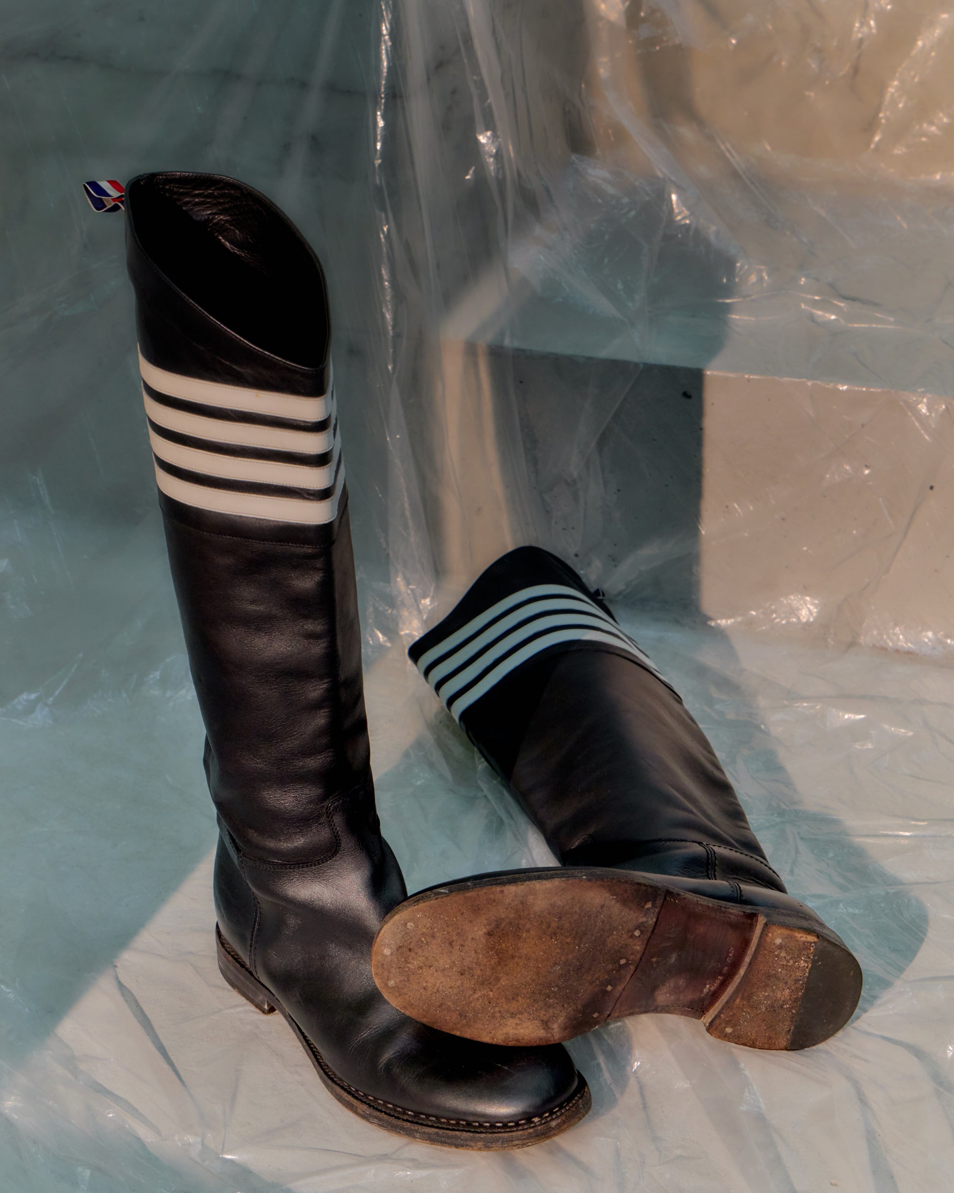 Pair of black Thom Browne riding boots with striped socks on a clear plastic sheet.