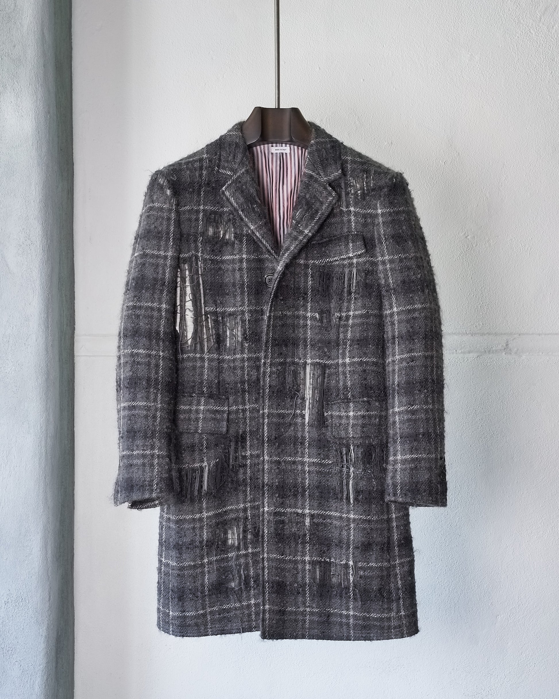 The Thom Browne FW 2016 Distressed Tartan Mohair Coat, featuring a grey plaid pattern with frayed patches and missing sections, hangs on a black hanger against a plain white wall.